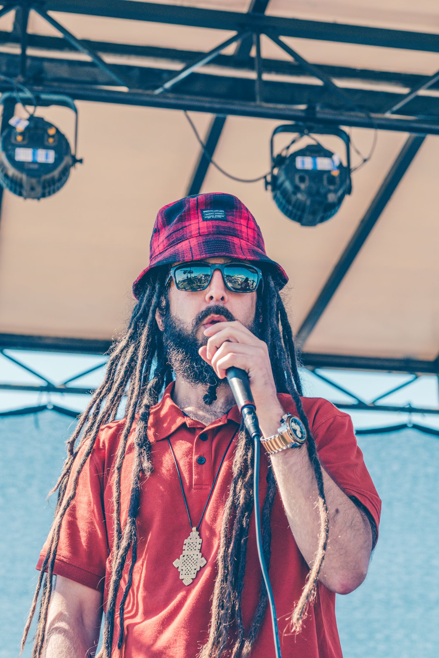 A bearded reggae artist with long dreadlocks and a plaid bucket hat sings into a microphone on stage.