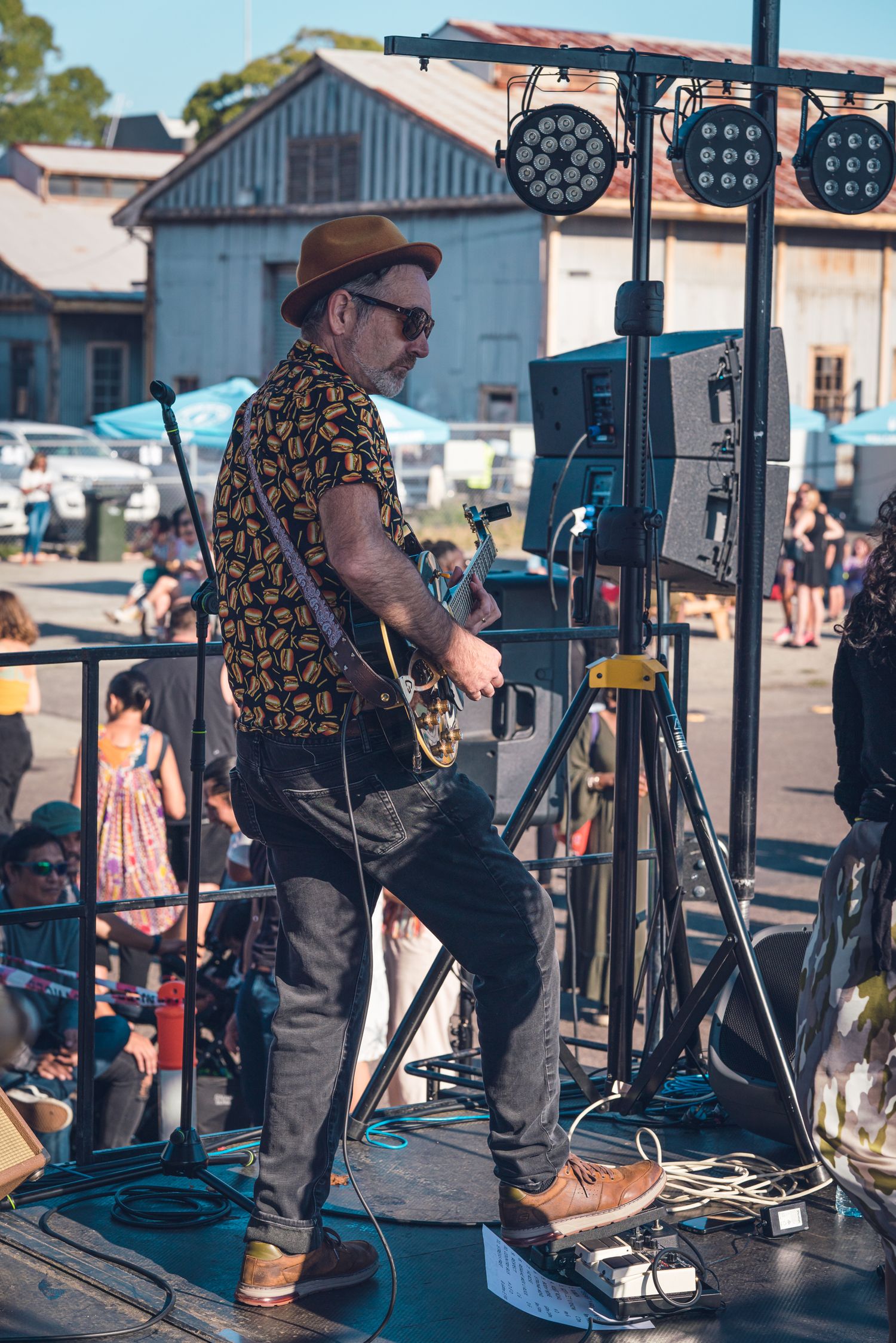 Male guitarist in floral shirt and brown hat performing on outdoor festival stage for crowd.