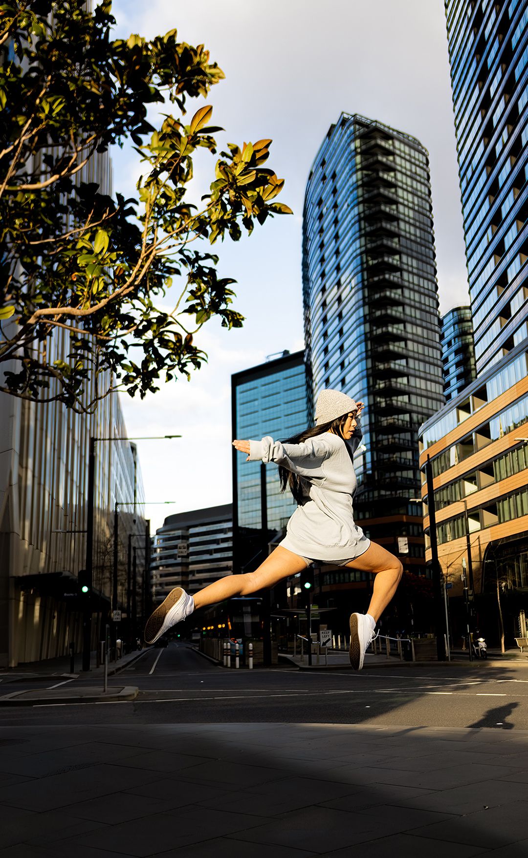 A woman leaps mid-air in a grey outfit on a city street surrounded by modern glass skyscrapers.