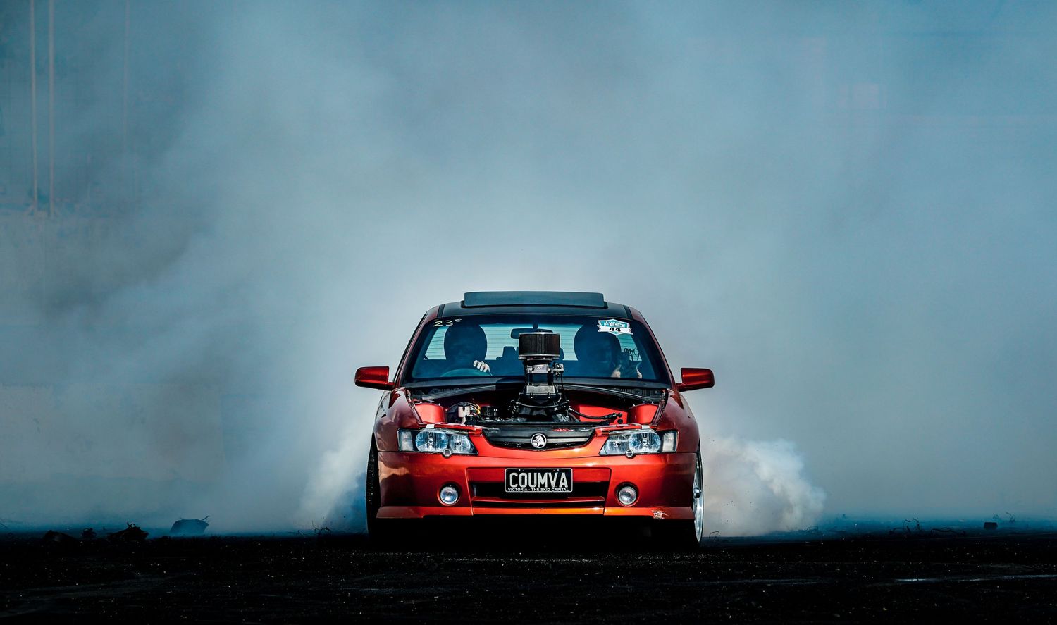 A red modified car emerges from thick white tire smoke during a dramatic burnout.