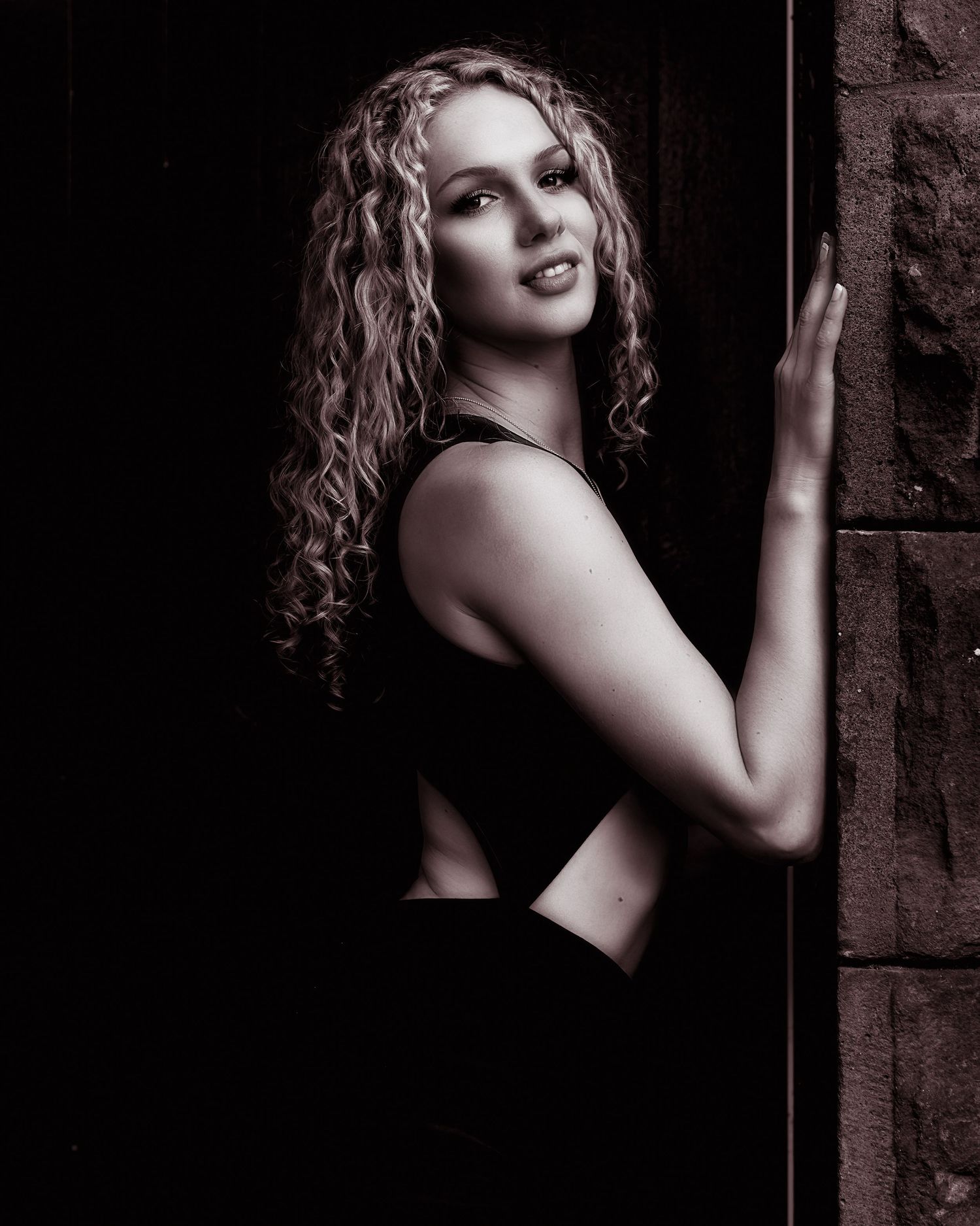 Young woman with curly blonde hair posing in black dress against dark stone wall in monochrome.