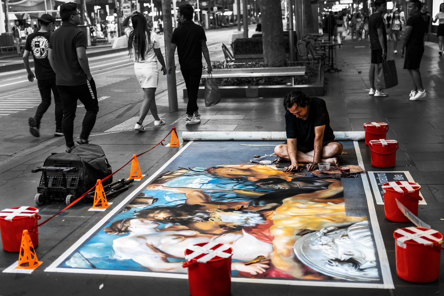 A street artist creates a detailed chalk painting on a busy pedestrian walkway in a city.