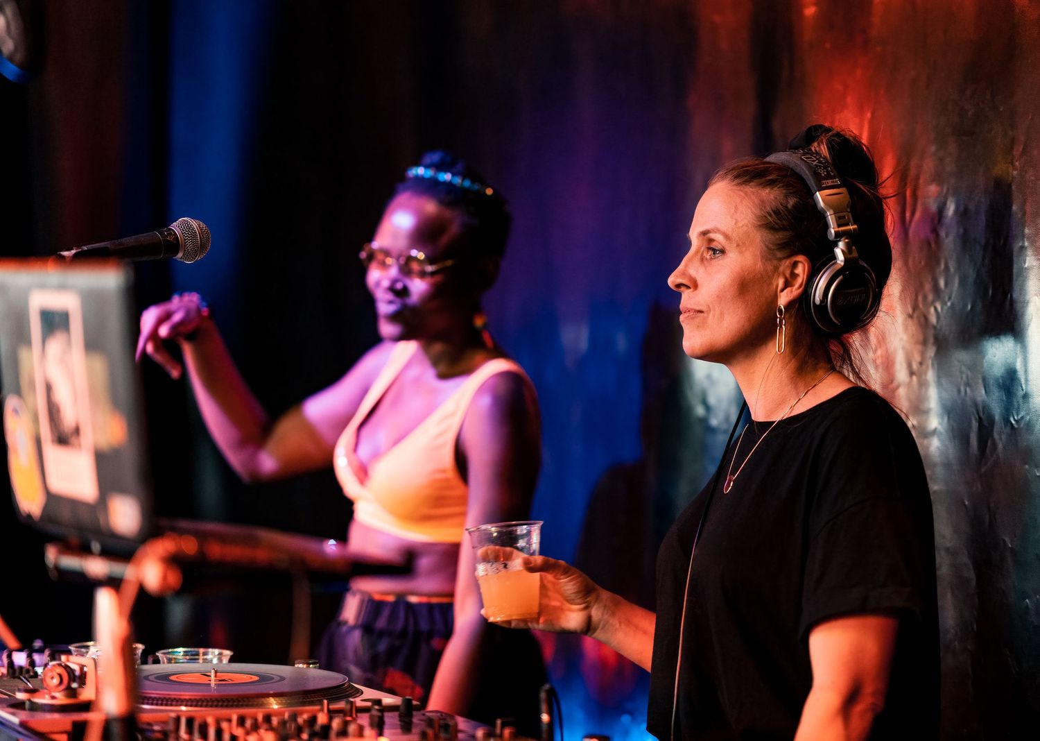 Two female DJs performing at a vibrant nightclub with colorful stage lighting and DJ equipment.