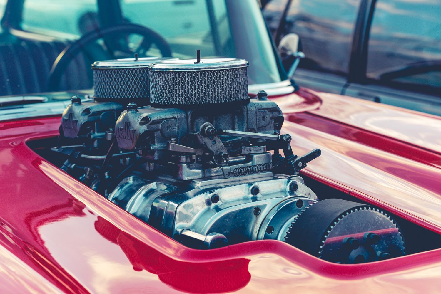 A chrome supercharged engine protrudes through the hood of a classic red car.