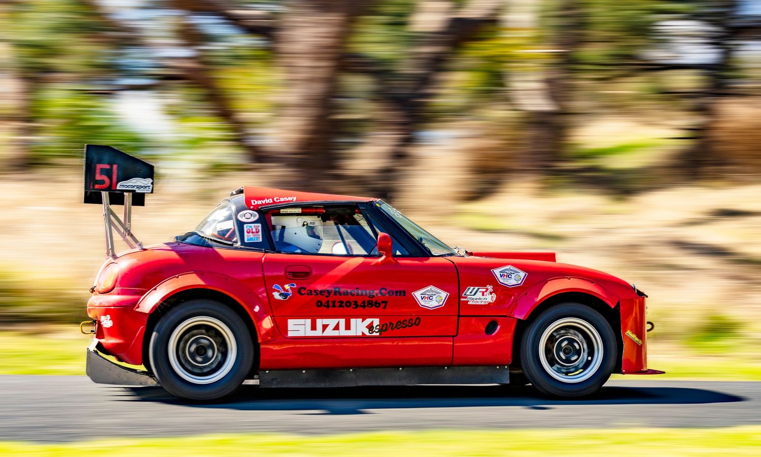 A red Suzuki race car races at high speed along a track with blurred trees in the background.