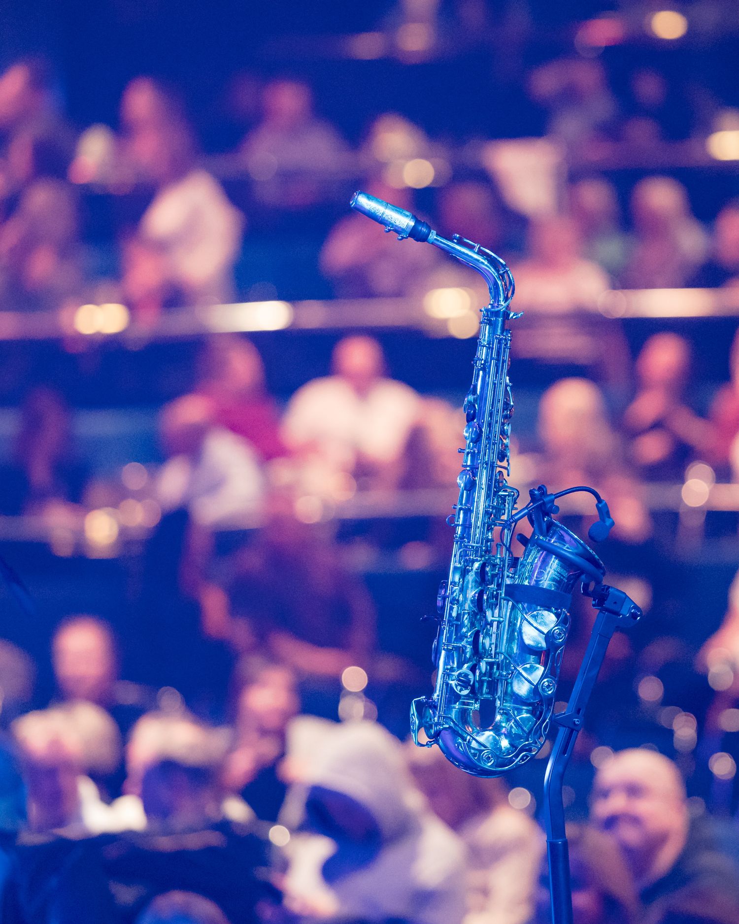 Alto saxophone bathed in blue stage lighting on a mic stand with a bokeh concert crowd in the background.