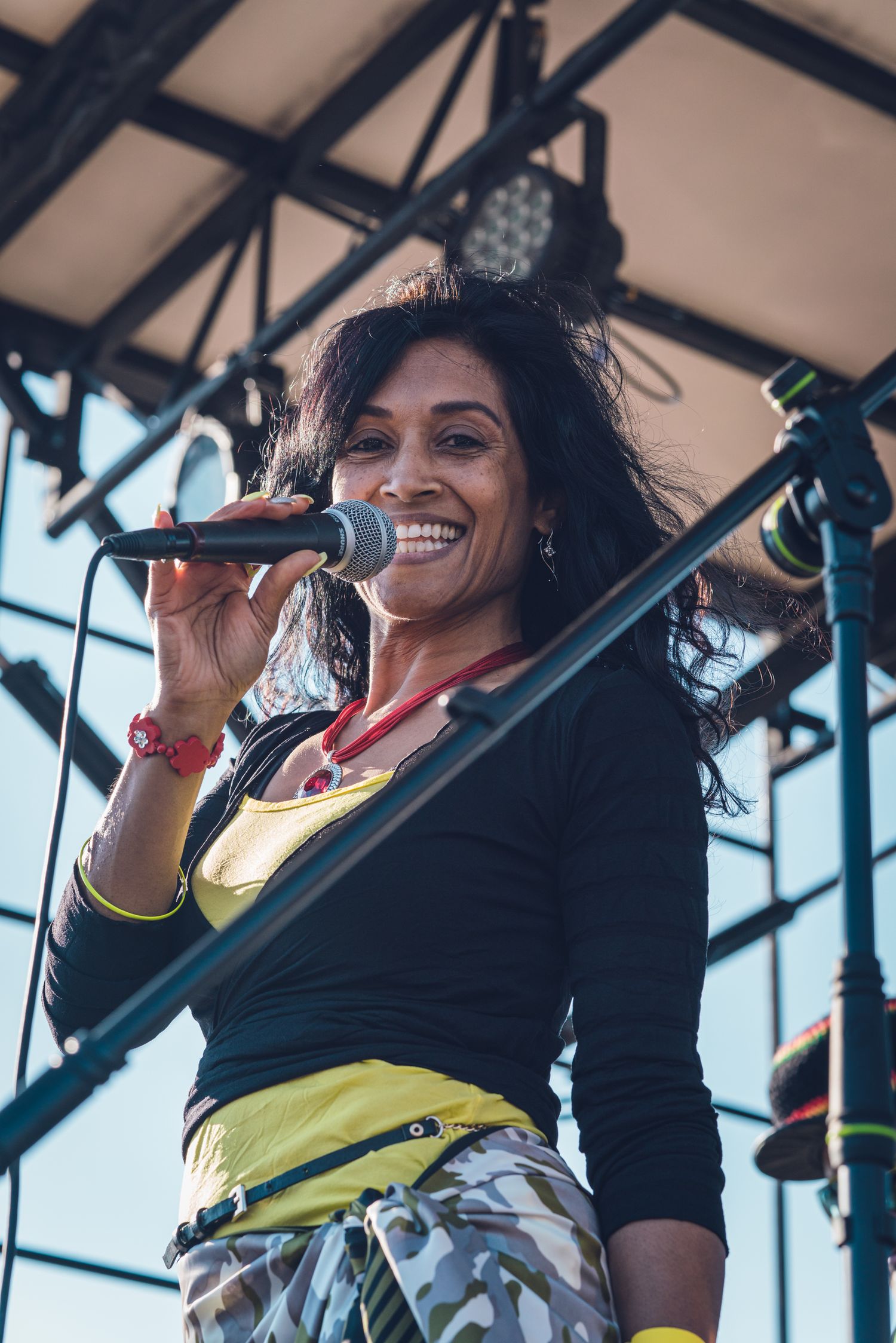 Smiling female vocalist in black and yellow top performing on outdoor festival stage.