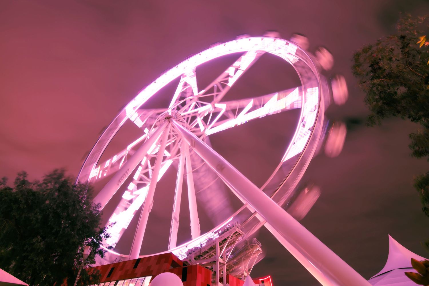 A glowing pink Ferris wheel spins against a dramatic night sky at an outdoor festival.