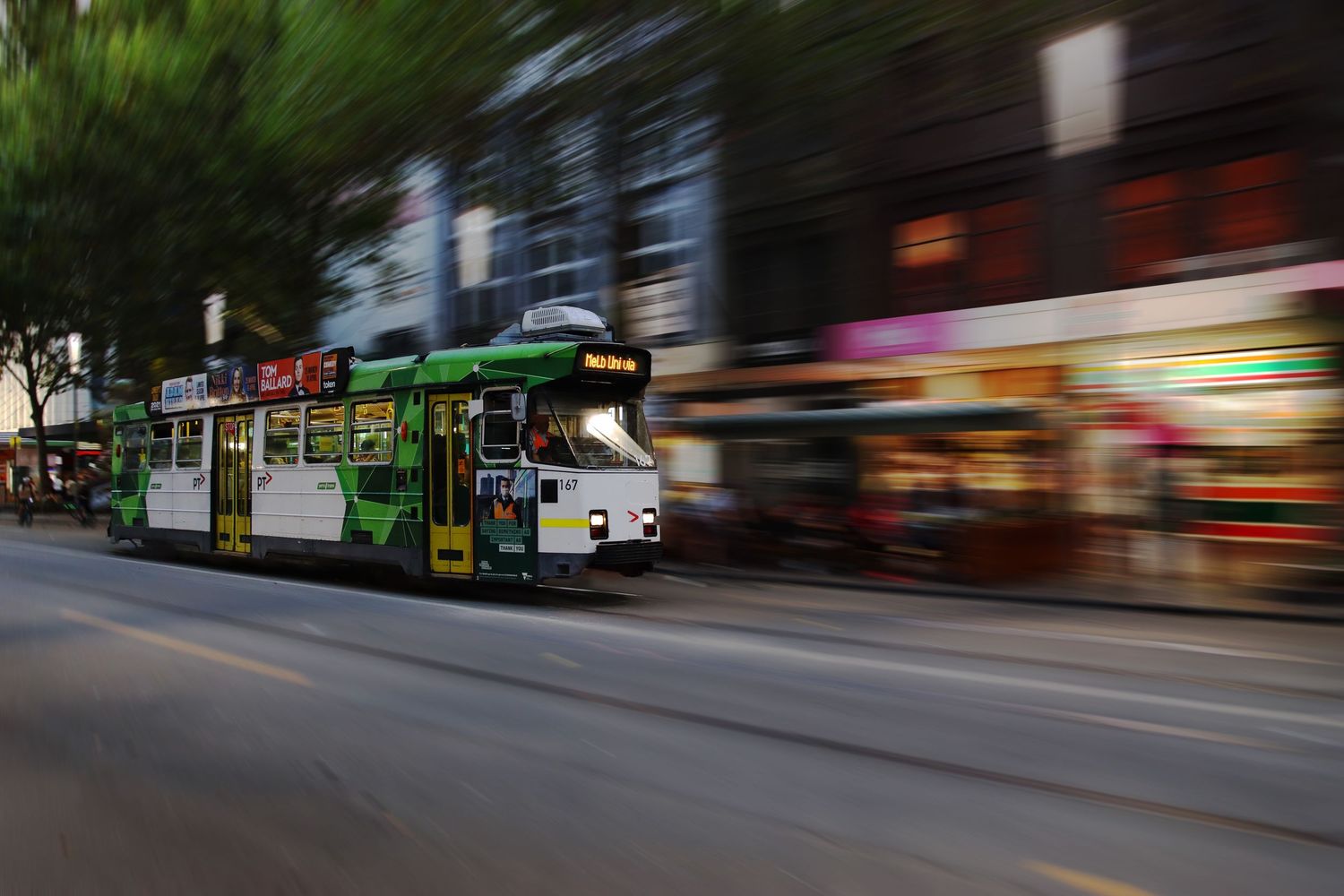 A green tram speeds through a city street at dusk, captured with motion blur photography.