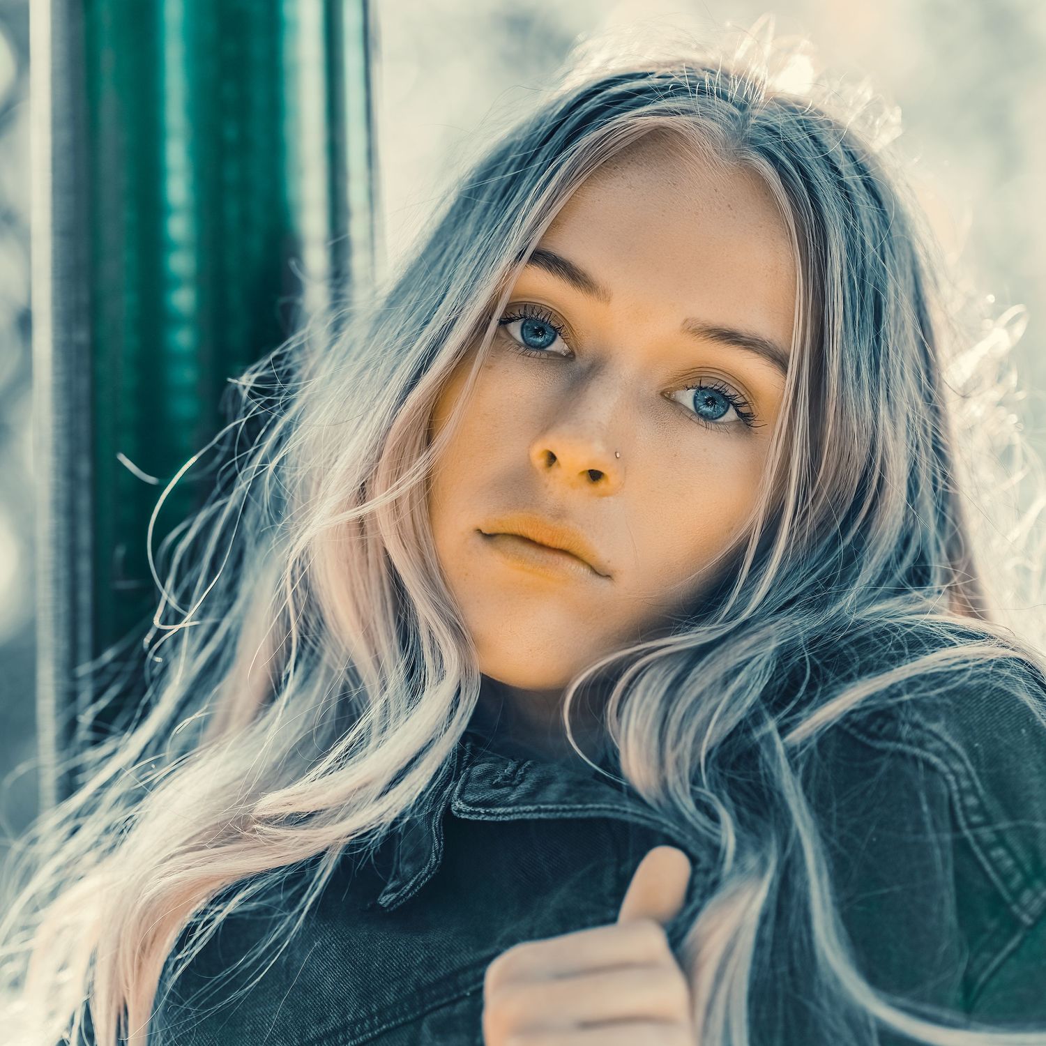 Close-up portrait of a young blonde woman with blue eyes wearing a denim jacket in soft natural light.
