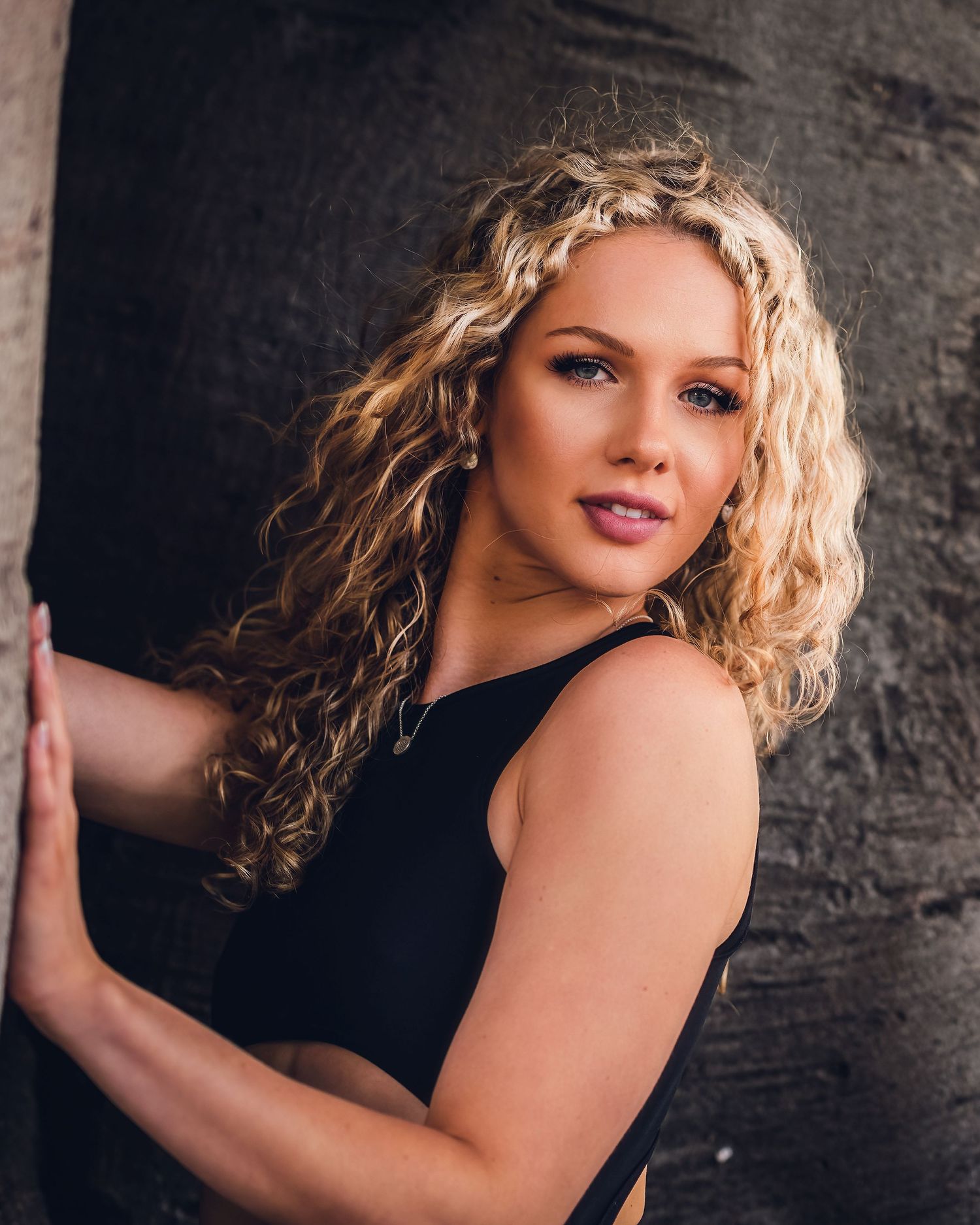 Professional portrait of a blonde curly-haired woman in a black top posing against a dark textured wall.