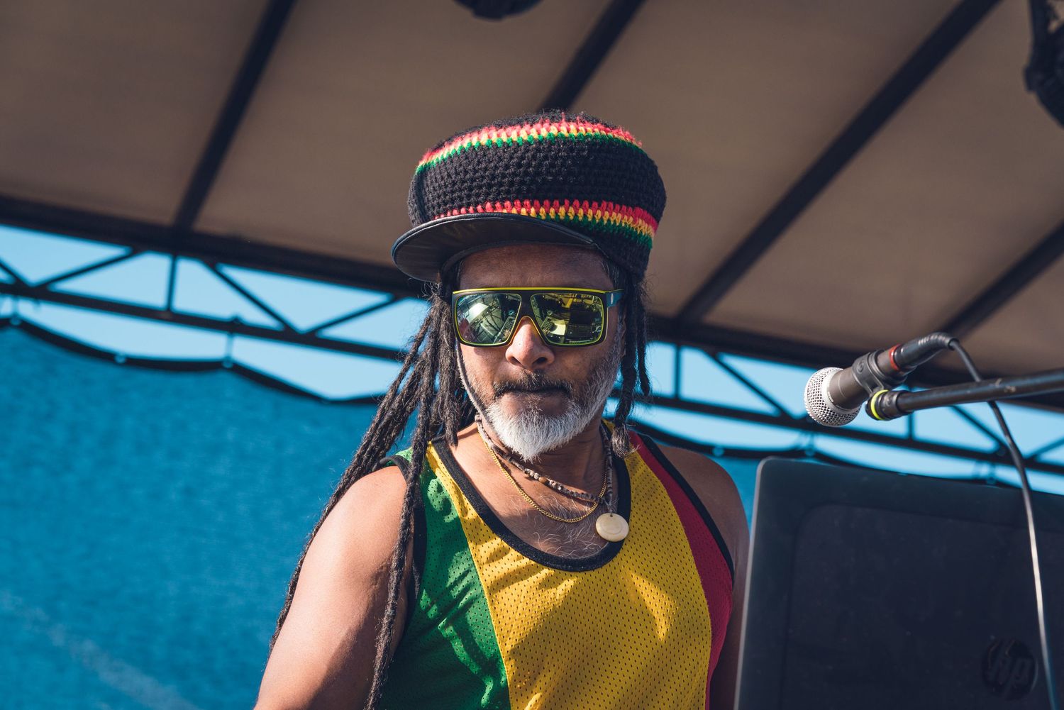 Rastafarian man wearing colorful knit hat and dreadlocks performing on stage at outdoor music festival.