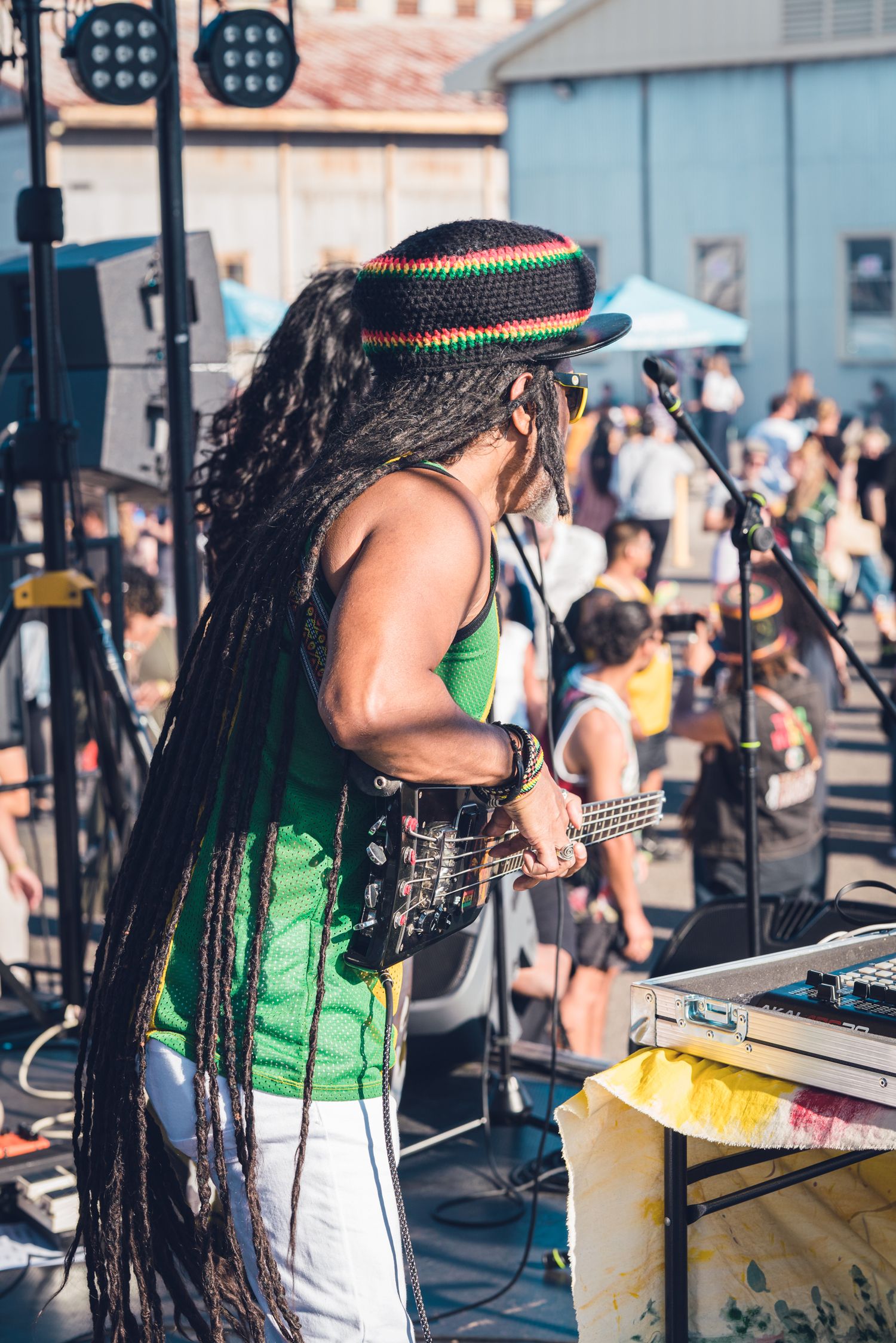 Reggae musician with long braids and Rasta hat playing guitar on outdoor festival stage.