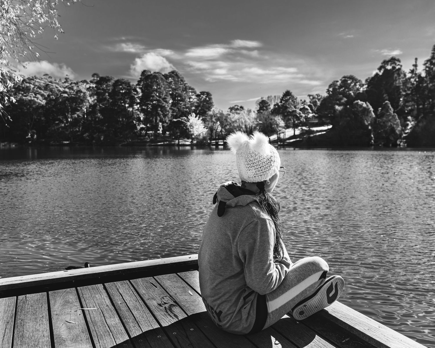 A woman in a white knit hat sits on a wooden dock, gazing at a serene lake in black and white.