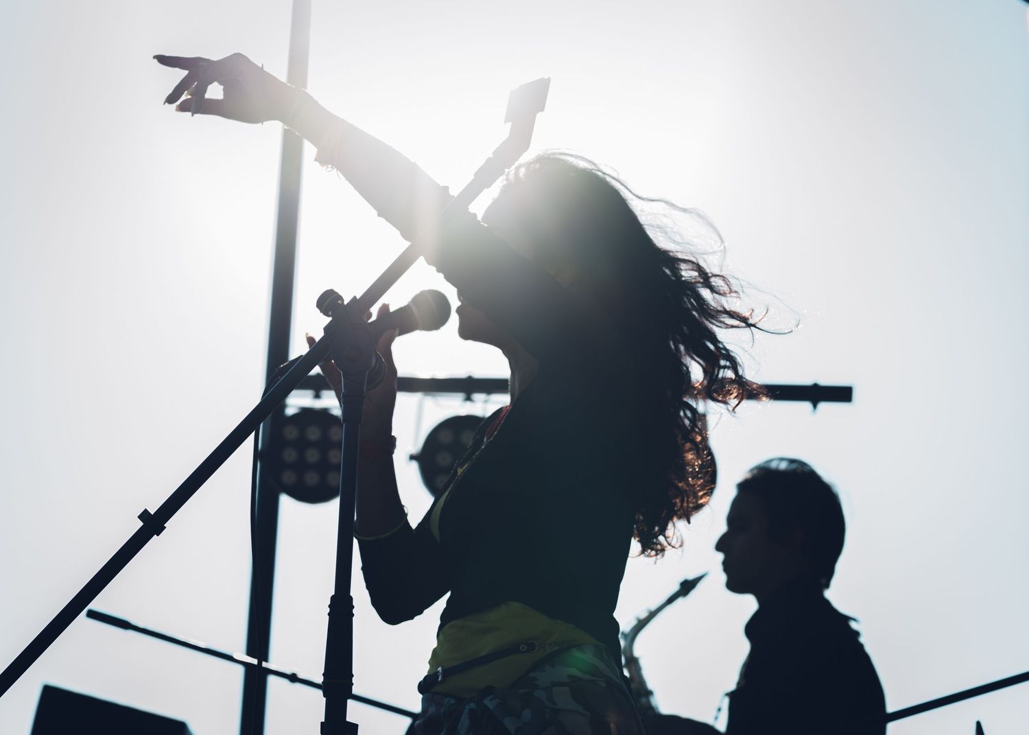 Dramatic silhouette of female singer raising arm while performing on stage against bright sunlit sky.
