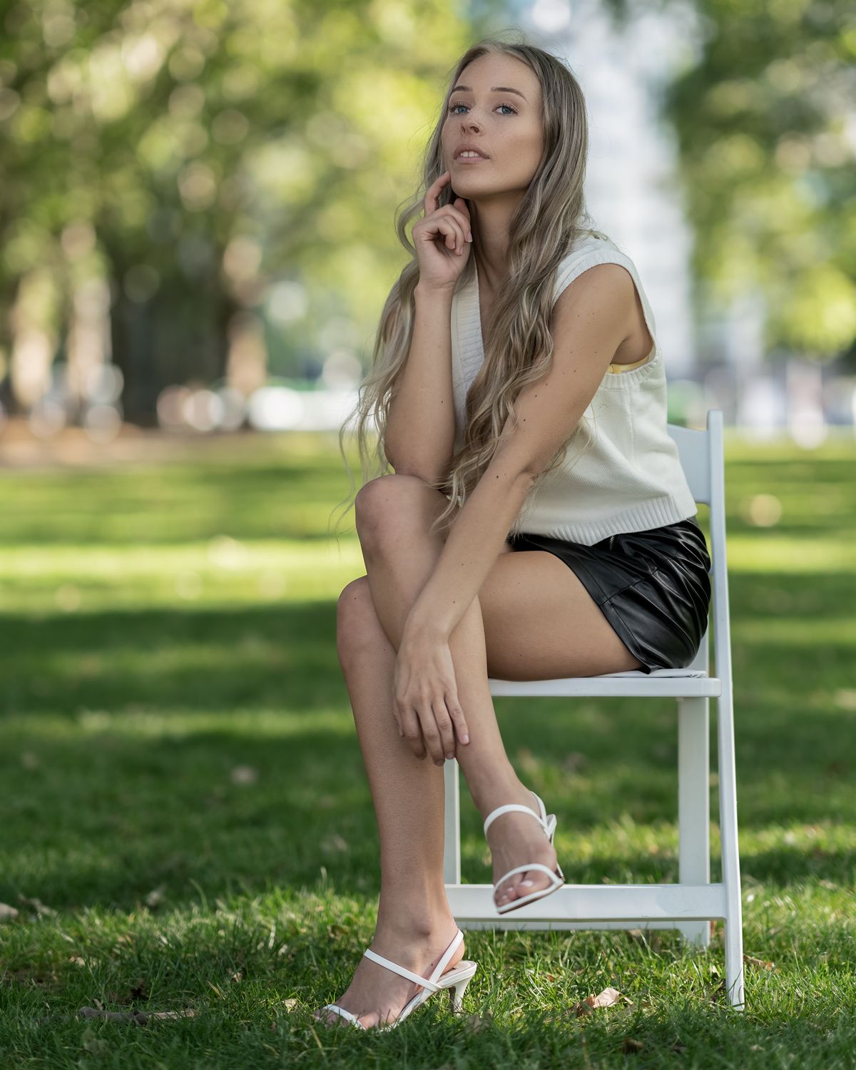Blonde woman in white top and black leather shorts sitting on a white chair in a sunny green park.
