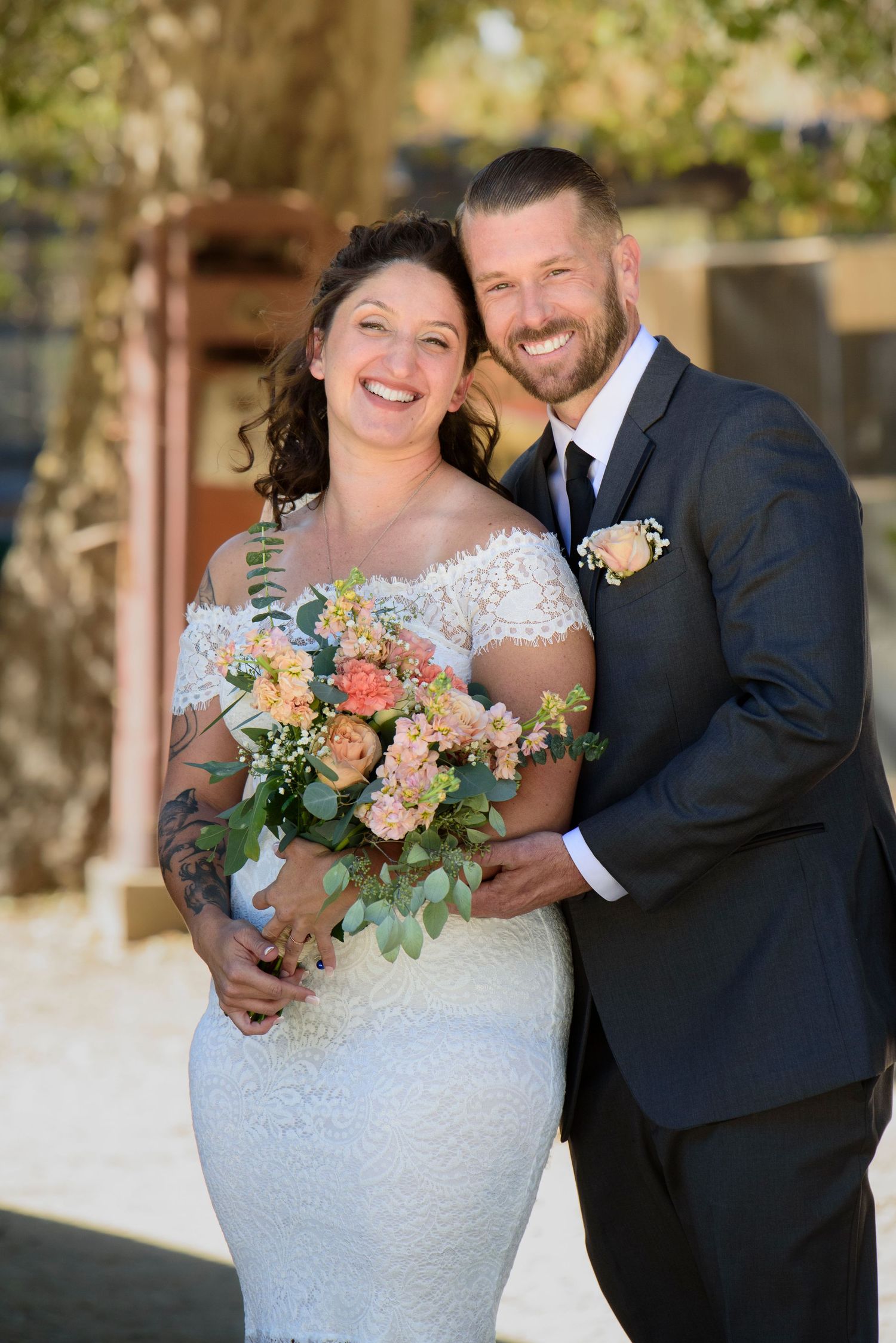 Smiling bride with tattoos holds peach floral bouquet as groom in dark suit embraces her outdoors.