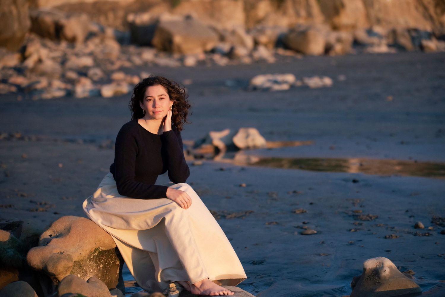 Young woman with curly hair sitting on rocks by the water in white skirt at sunset.