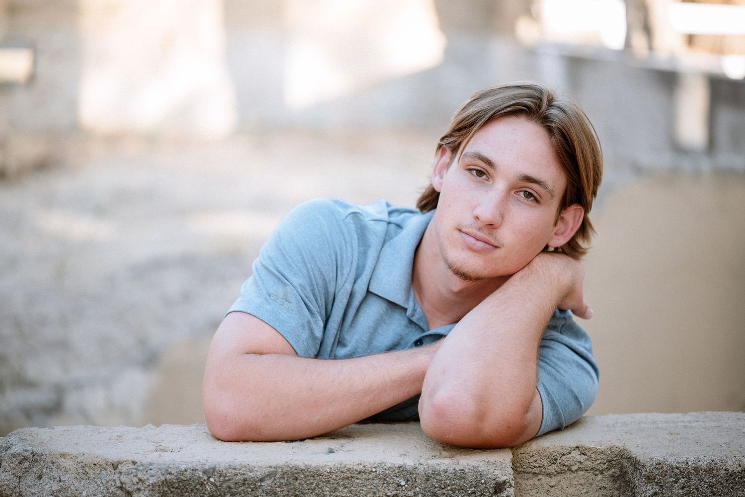 Young man with blonde hair rests chin on arms while leaning on concrete surface outdoors.