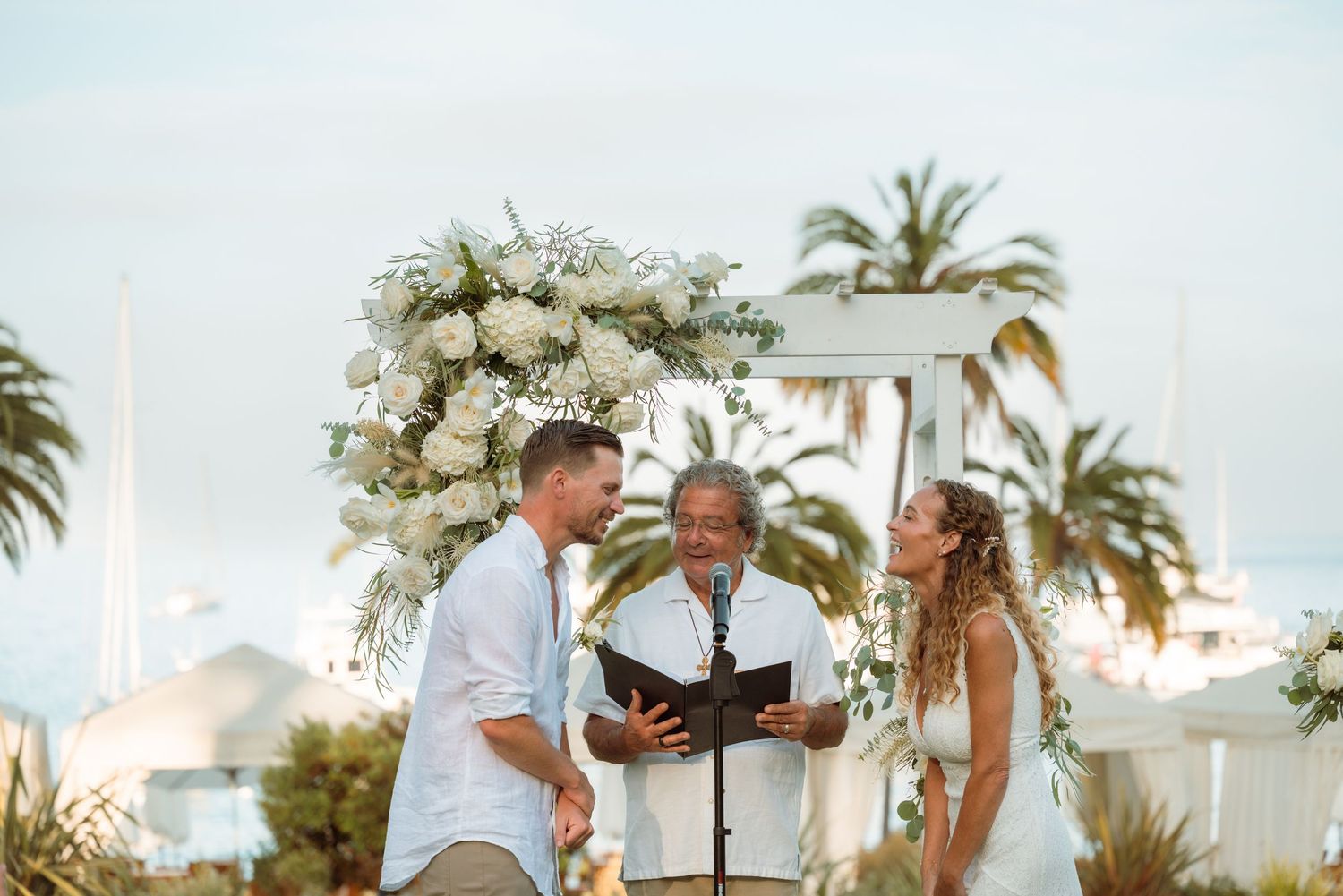 Couple exchanging vows at outdoor beach wedding ceremony under white floral arch with palm trees in background.