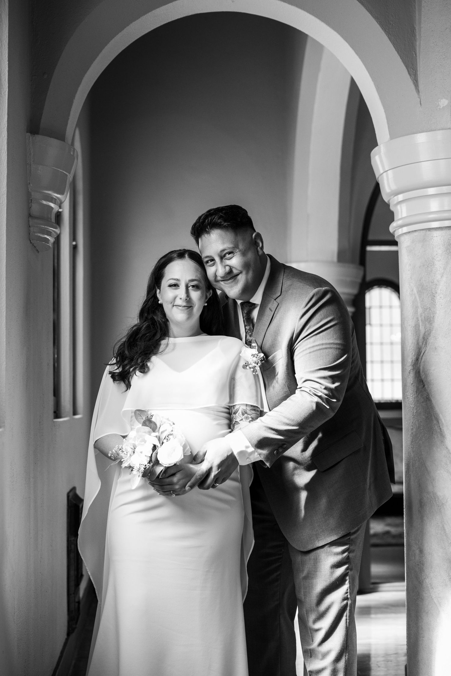 Black and white photo of newlyweds posing under elegant arches, bride holding bouquet in white gown, groom in suit smiling.