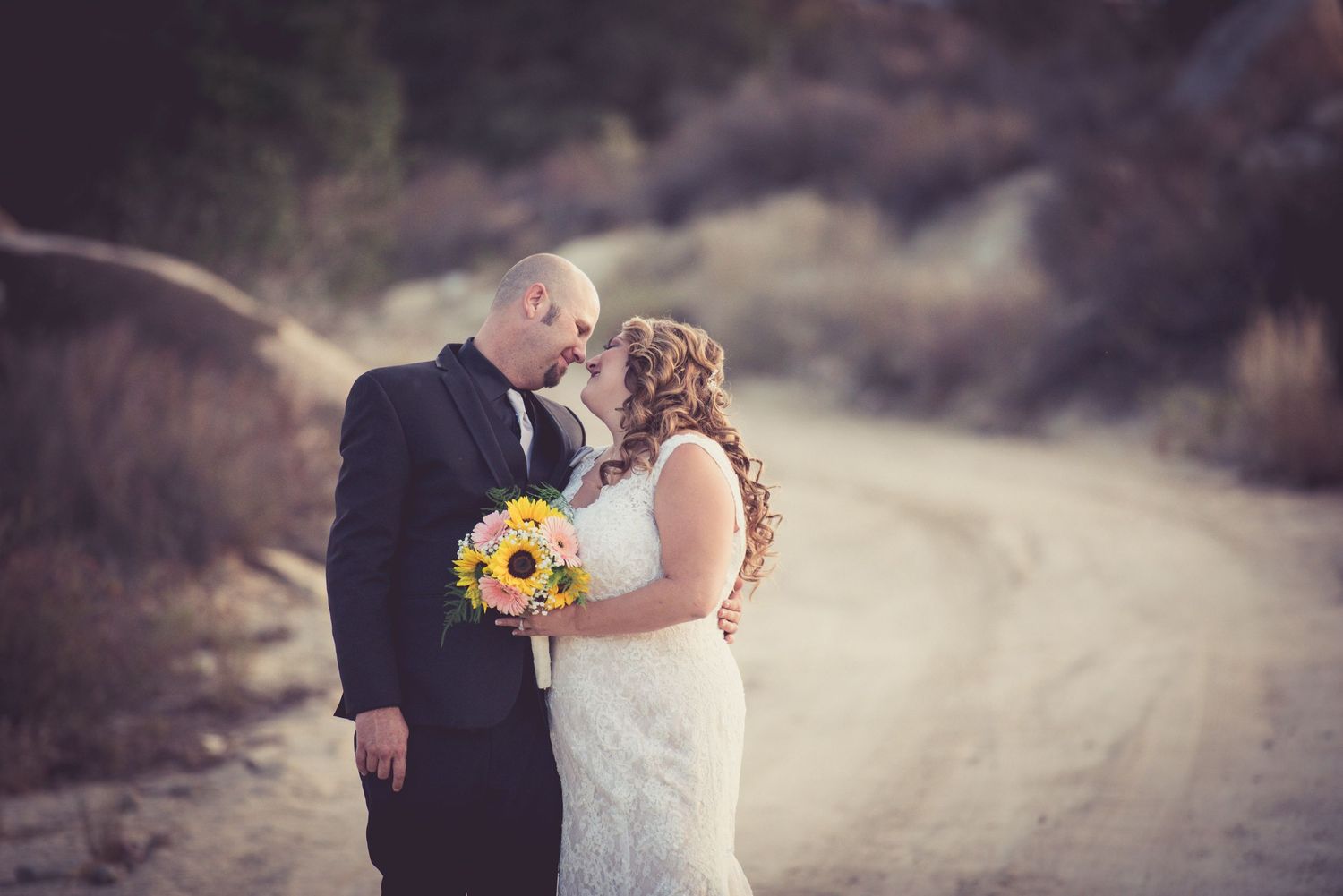 Bride and groom sharing kiss on dirt road as bride holds vibrant sunflower bouquet in desert setting.