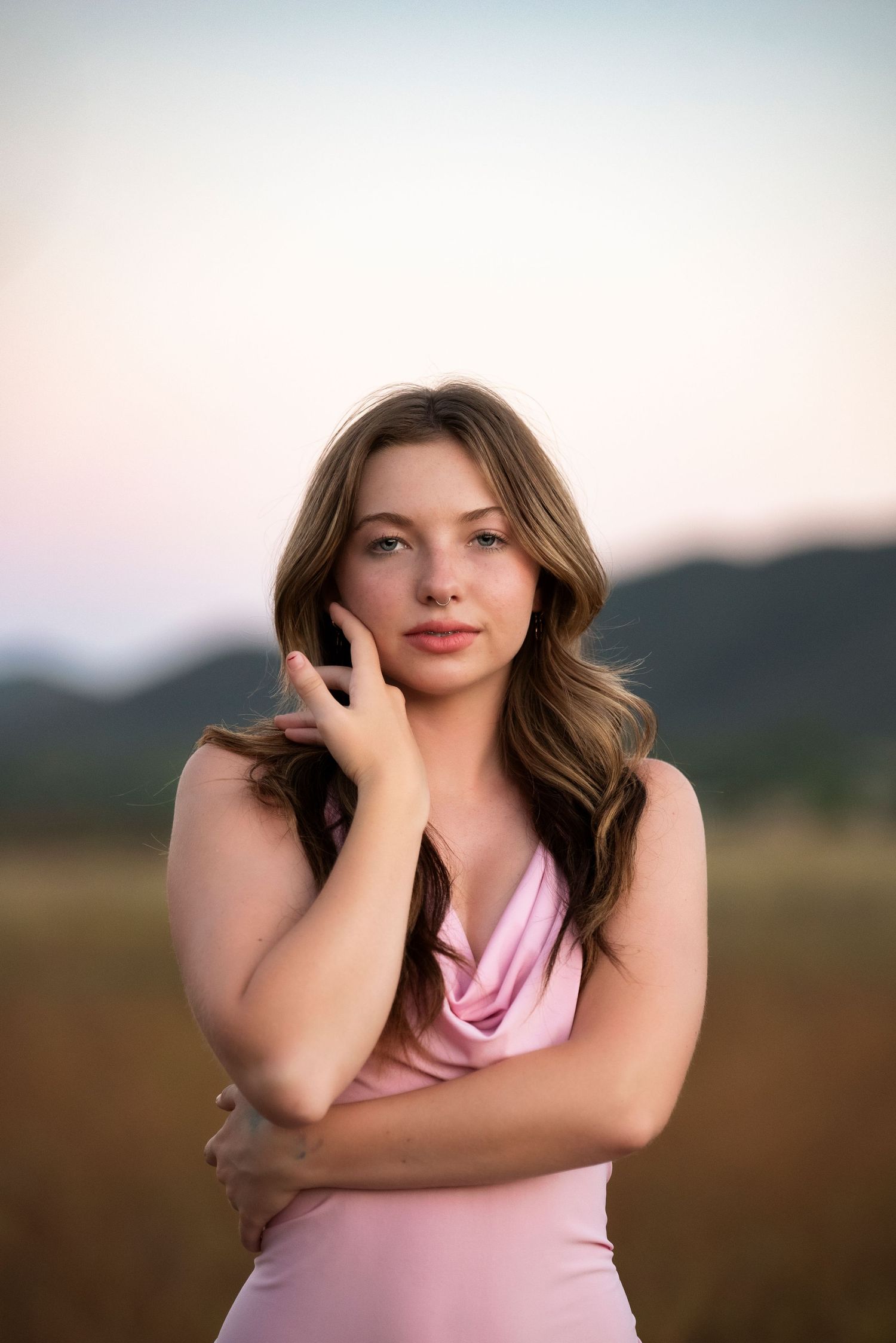 Young woman in pink cowl neck dress posing thoughtfully in an open field at dusk with mountains.