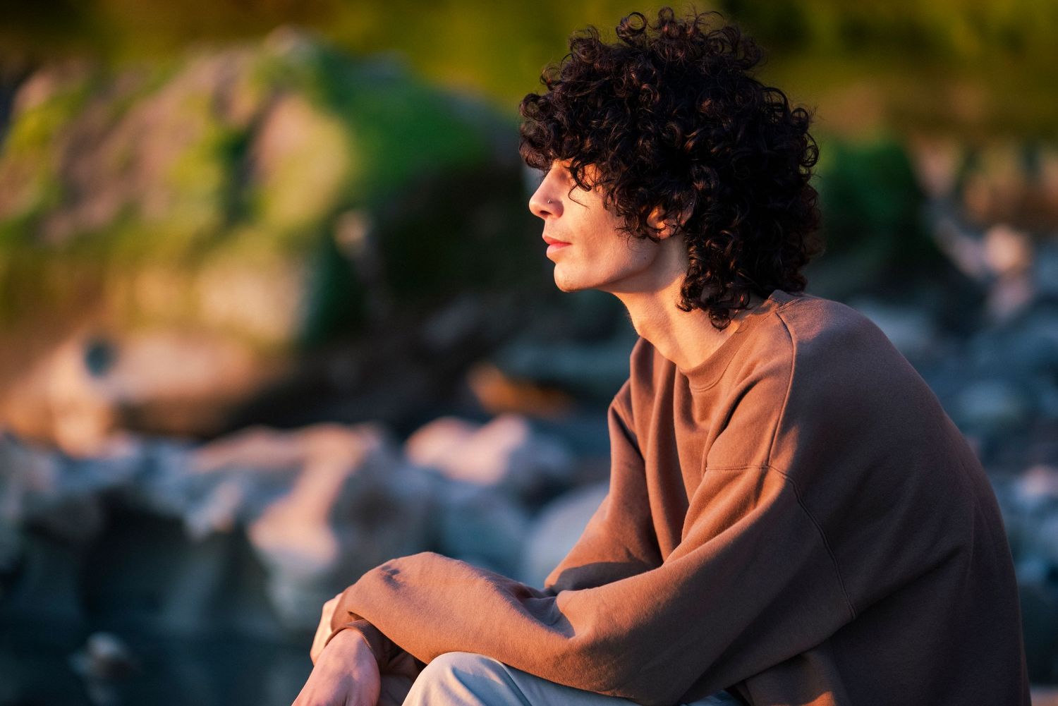 Young man with curly hair sitting on rocks gazing into distance in warm sunset light.