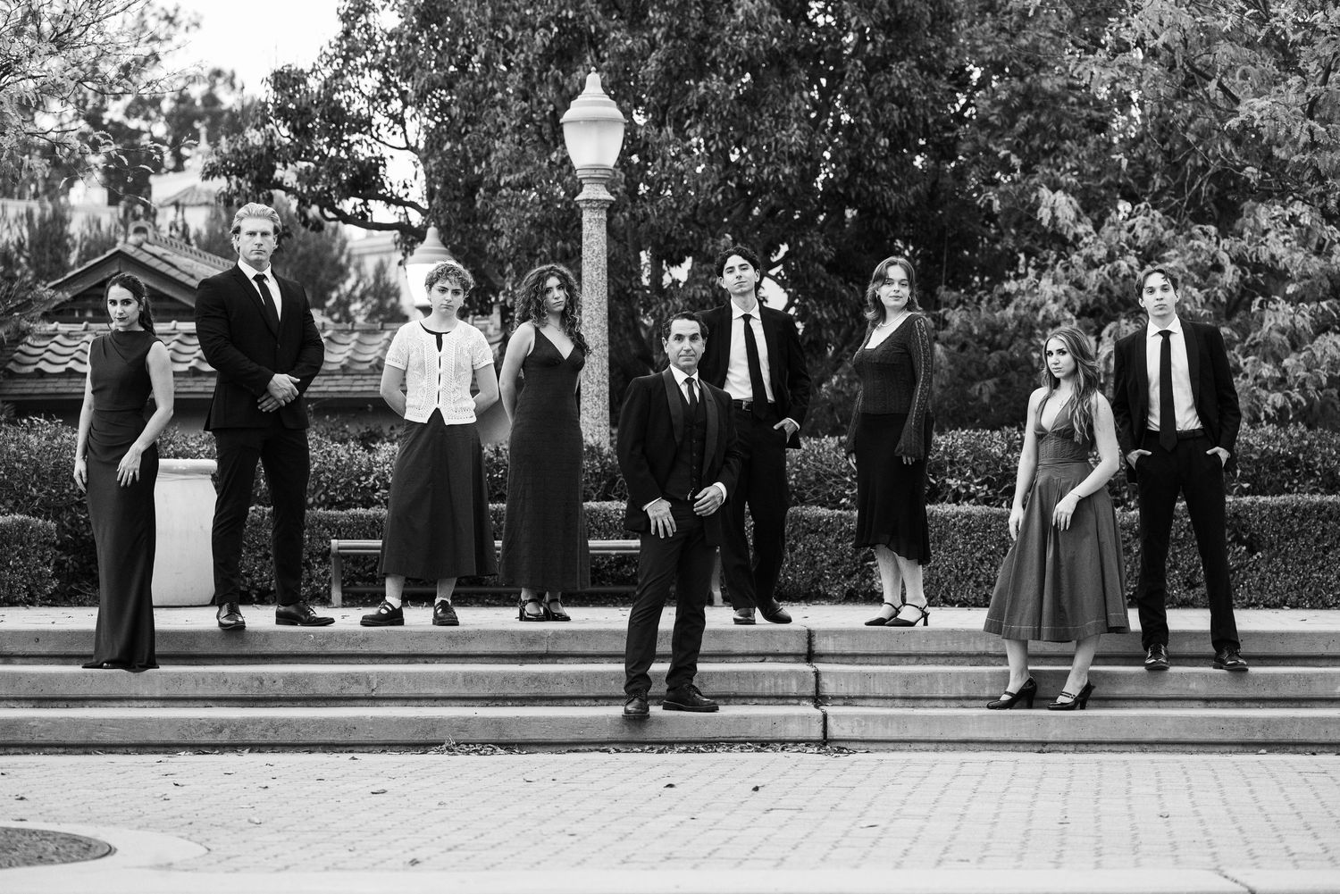 Black and white photo of eight formally dressed men and women posing on outdoor steps with trees and a lamppost behind them.