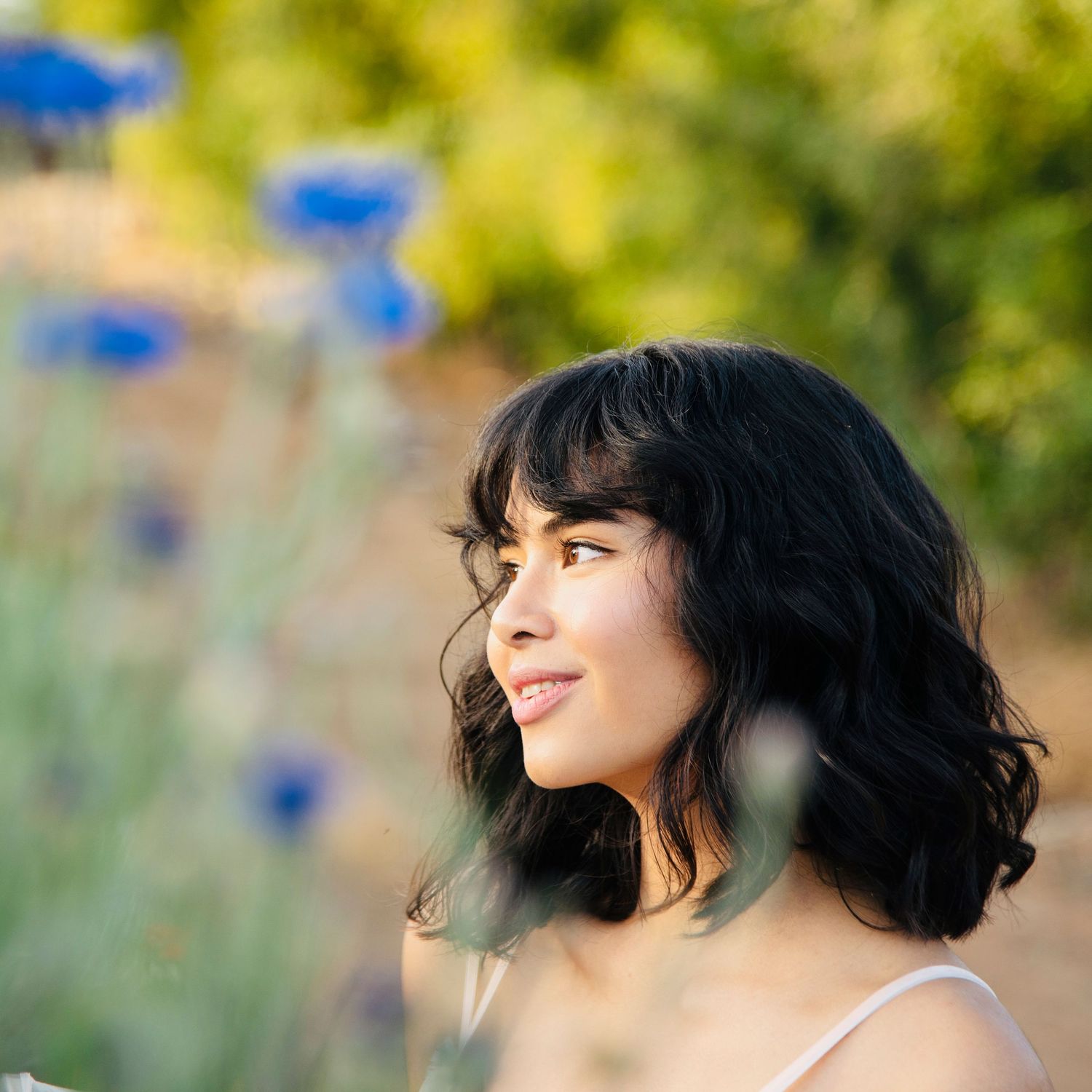Young woman with short dark wavy hair smiling softly outdoors with blue wildflowers in background.