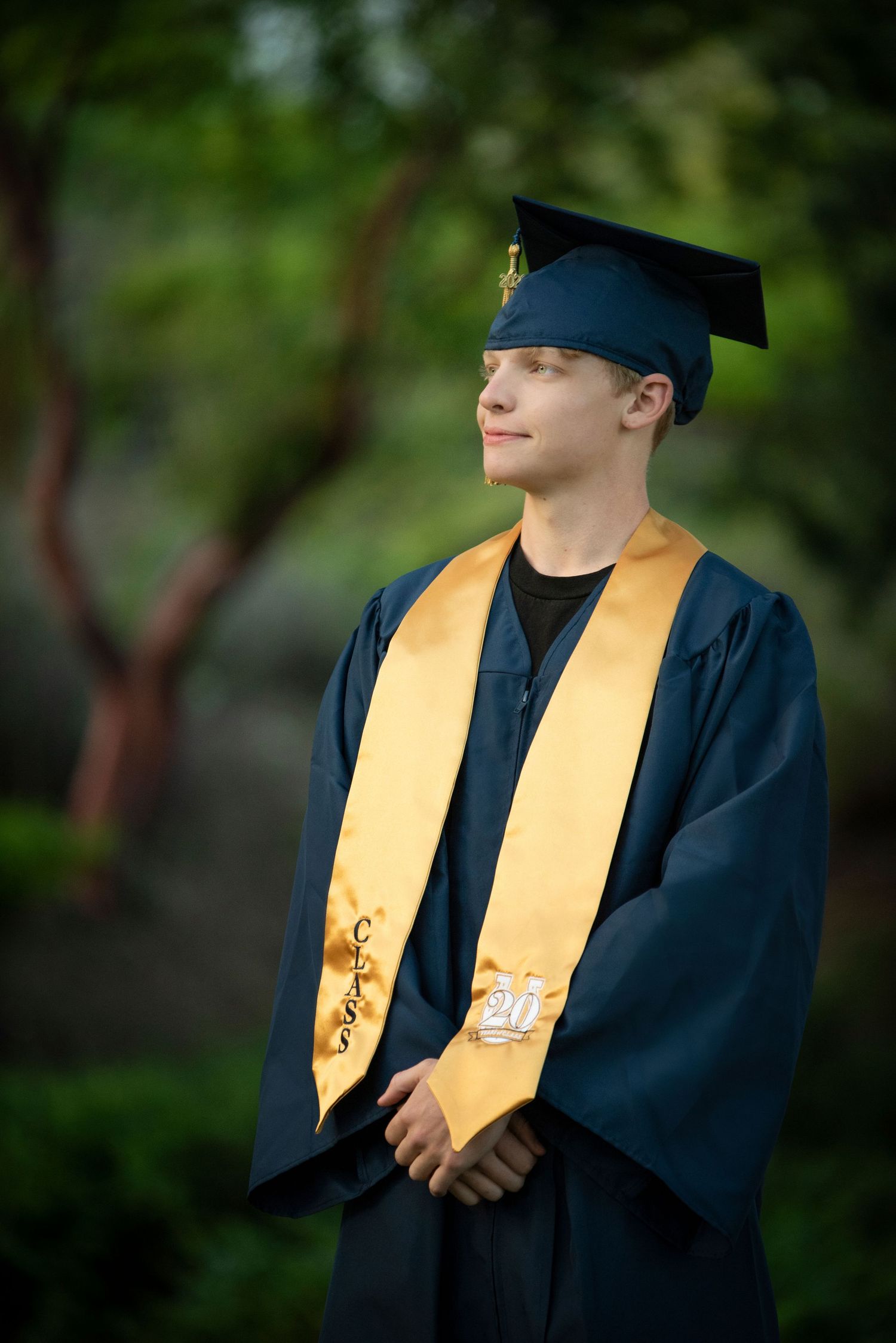 Young man in navy graduation cap and gown with gold honor stole smiling outdoors among green trees.