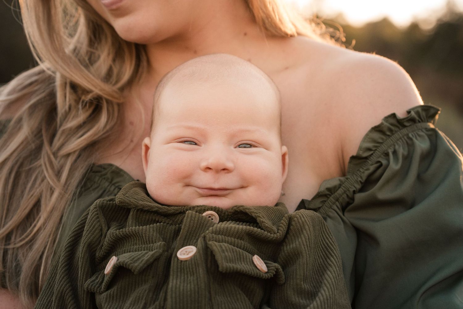 Adorable smiling baby in green outfit being cradled by mother in warm golden light.