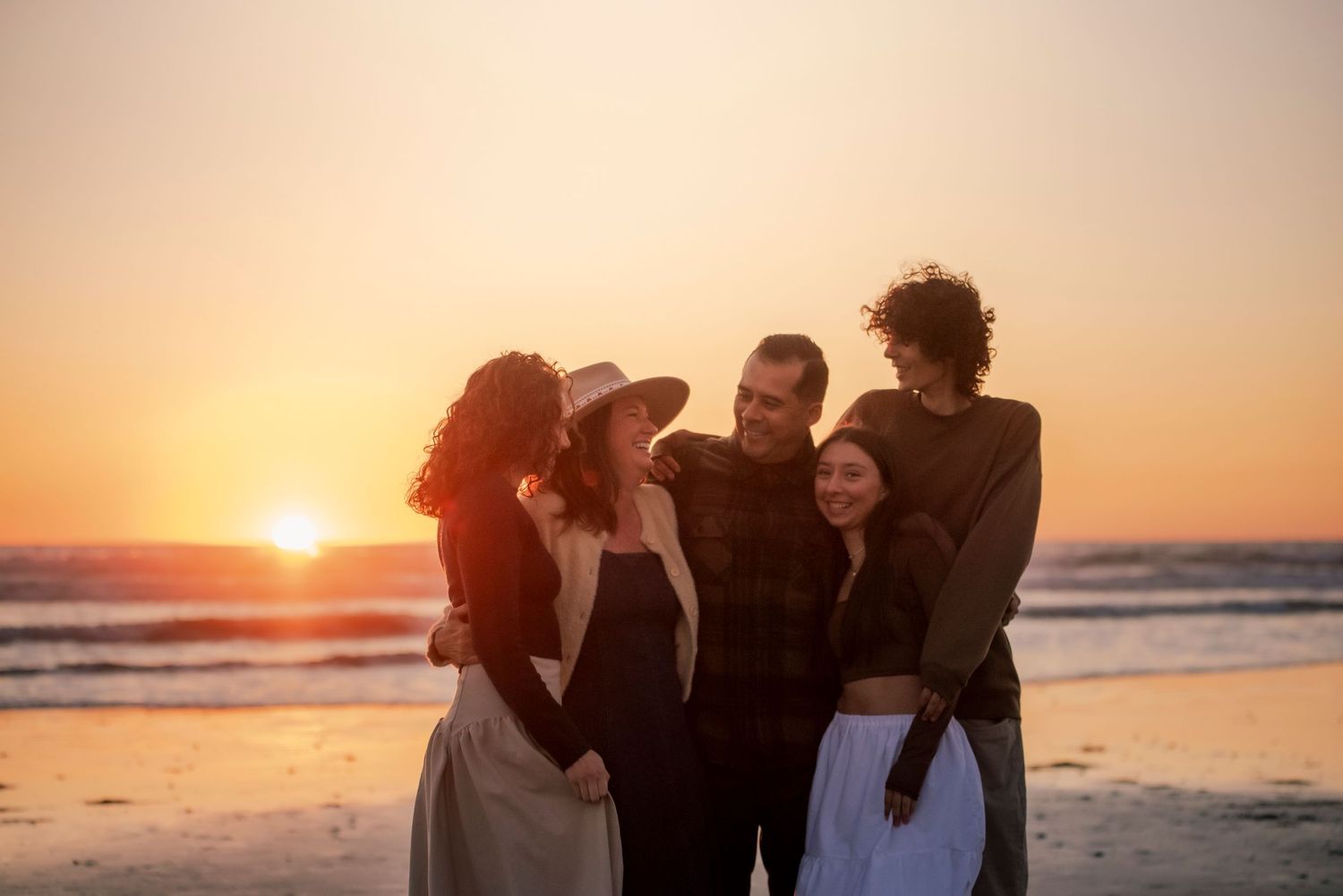 A joyful family of five embraces together during a stunning golden sunset at the beach.