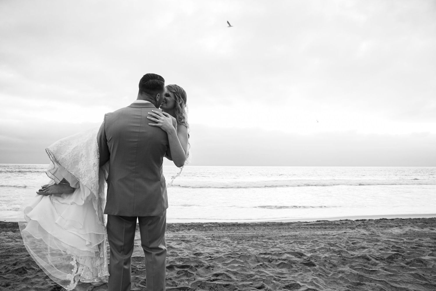 Black and white photo of newlywed couple standing together on a beach, looking at the ocean.