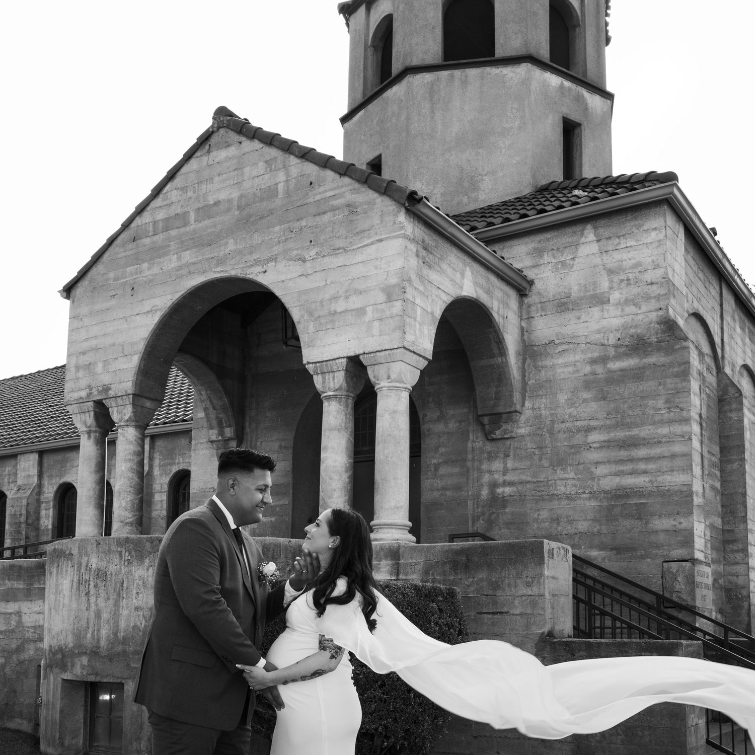 Black and white wedding photo of couple embracing before a historic stone church with a bell tower.