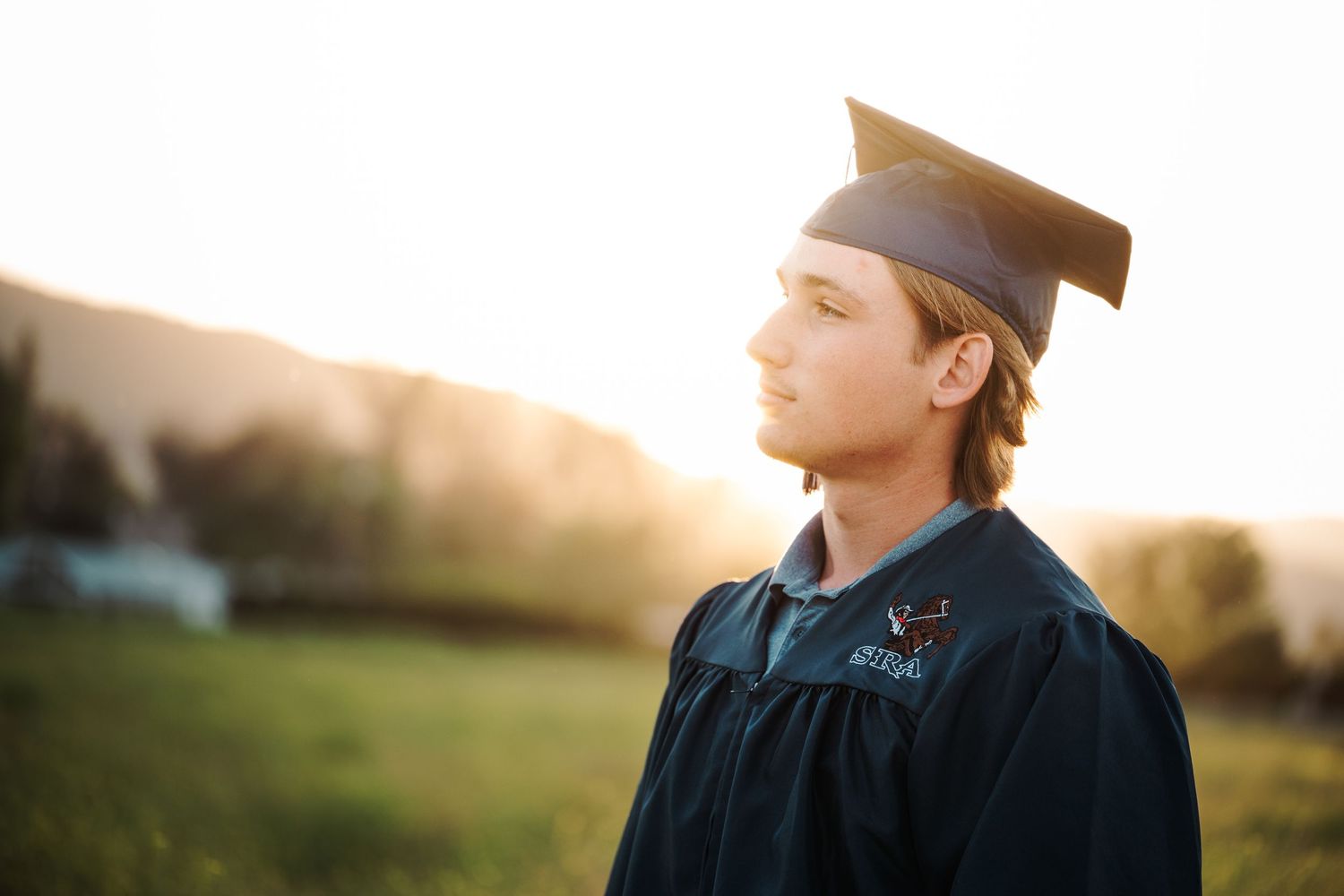 Graduate in blue cap and gown gazes into distance during golden sunset in green landscape.
