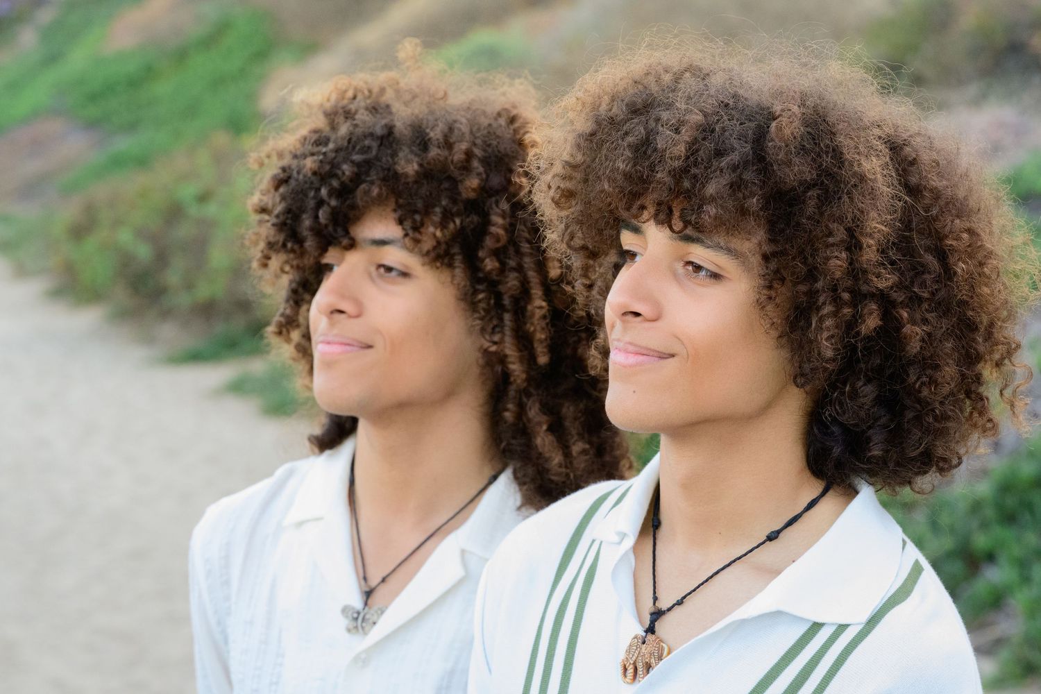 Two young male twins with curly hair wearing matching white shirts smile outdoors on a sunny day.