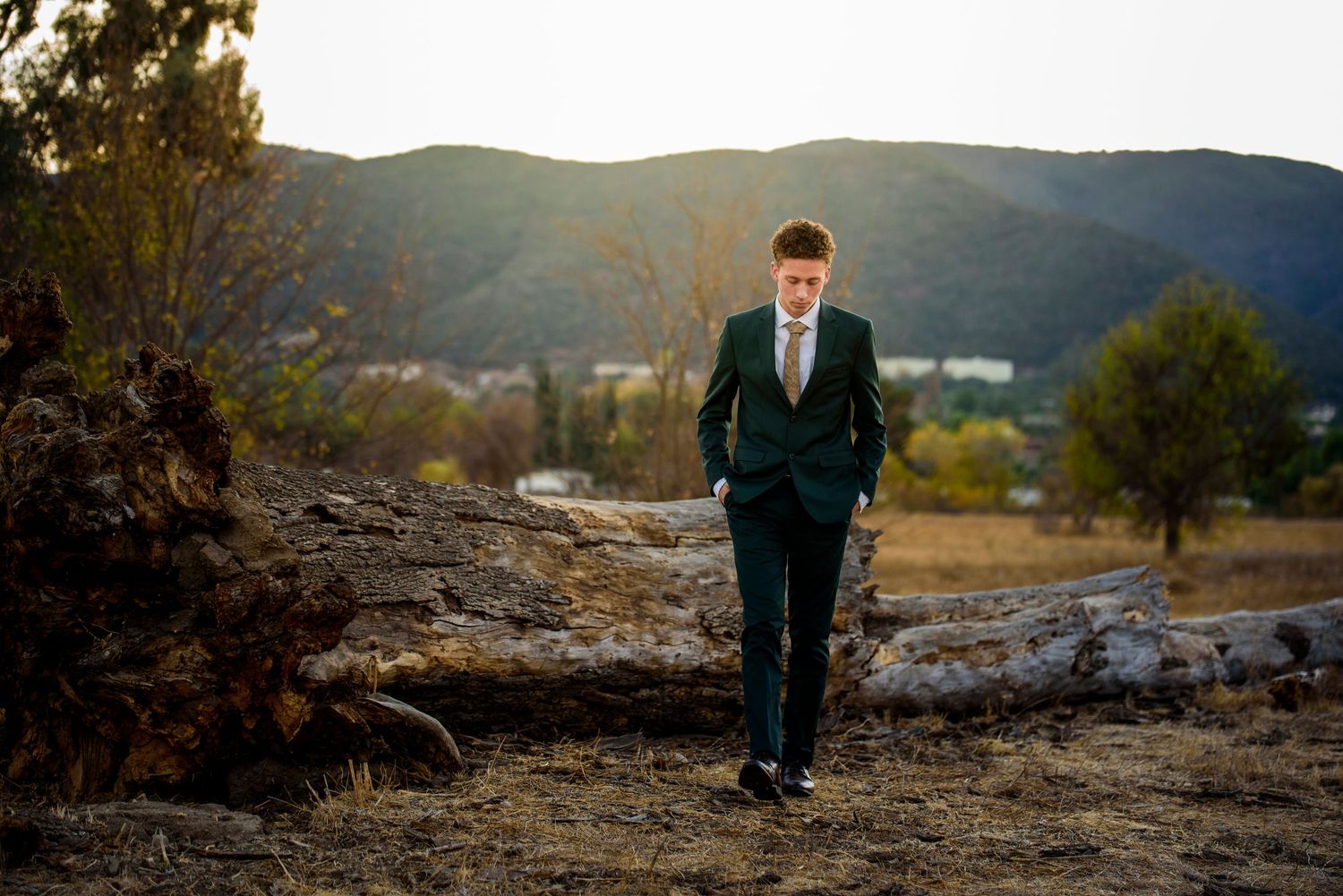 Young man in dark suit walking outdoors at golden hour with mountain landscape in background.