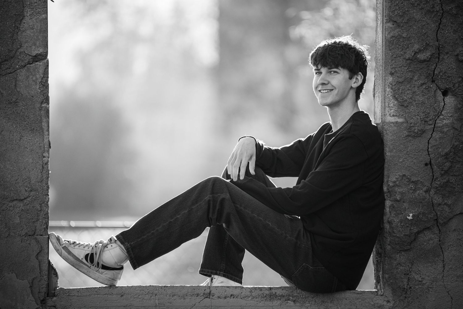 Smiling young man in black outfit sitting relaxed in stone window frame in black and white photo.