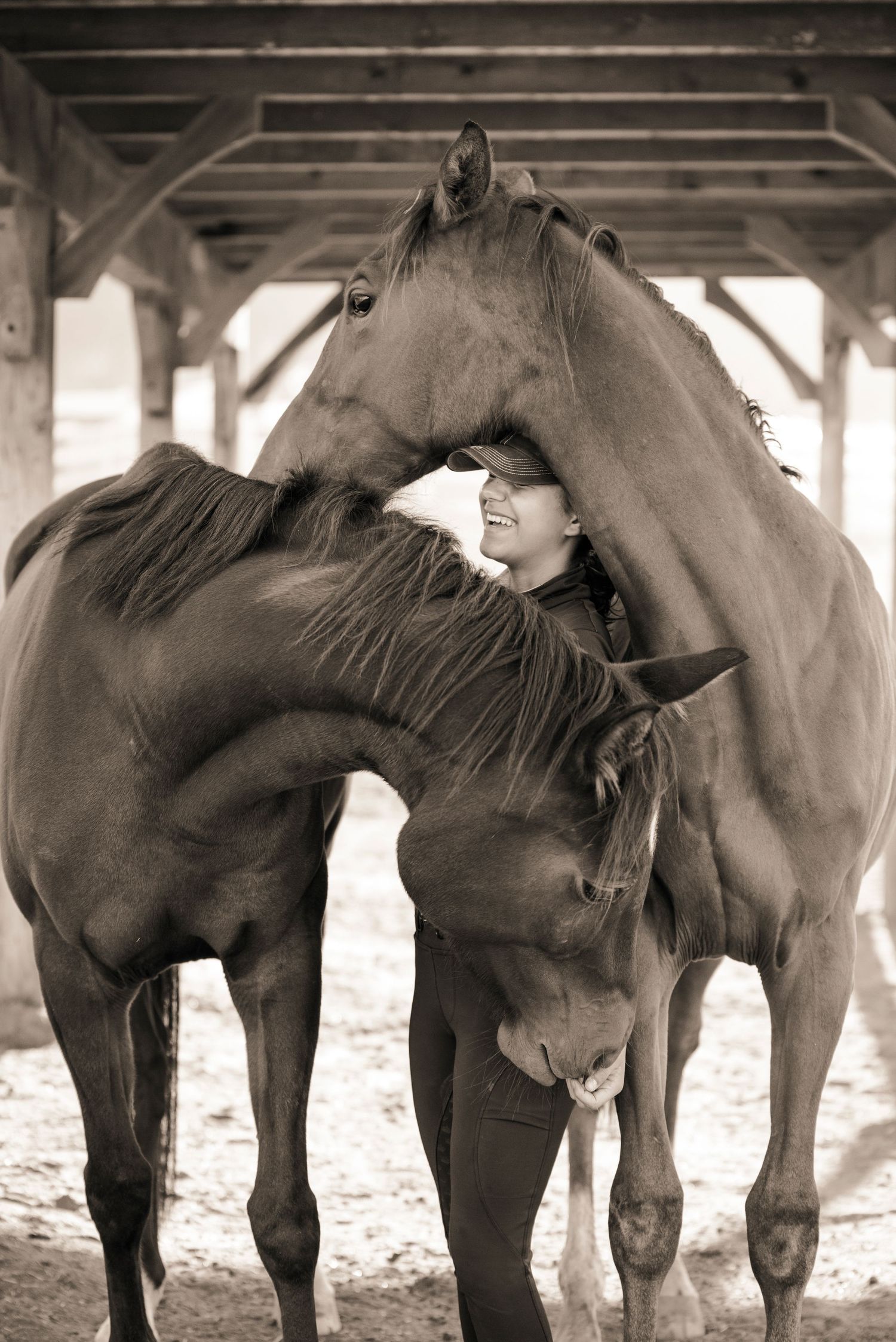 Sepia toned photo of a smiling woman surrounded by two affectionate horses under a stable shelter.