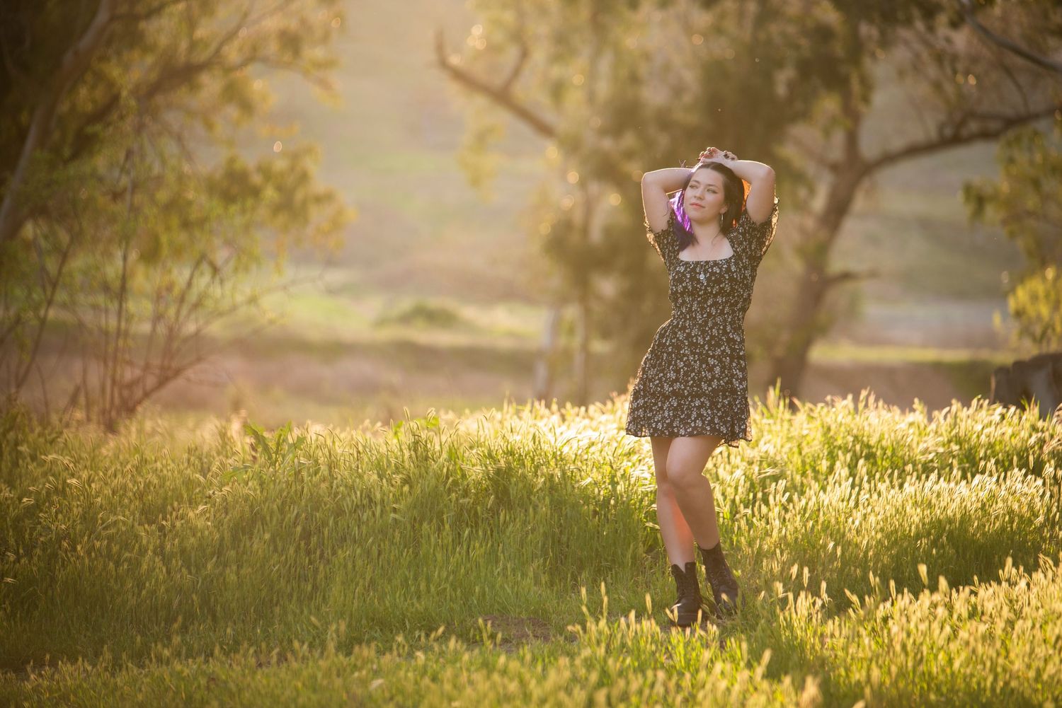 Young woman in floral dress and boots posing in a sunlit green meadow with trees.