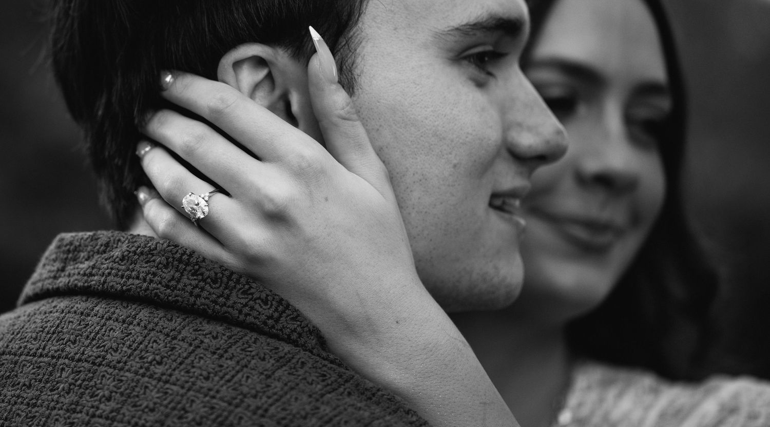 Black and white photo of a couple embracing, woman showing off a diamond engagement ring while smiling.