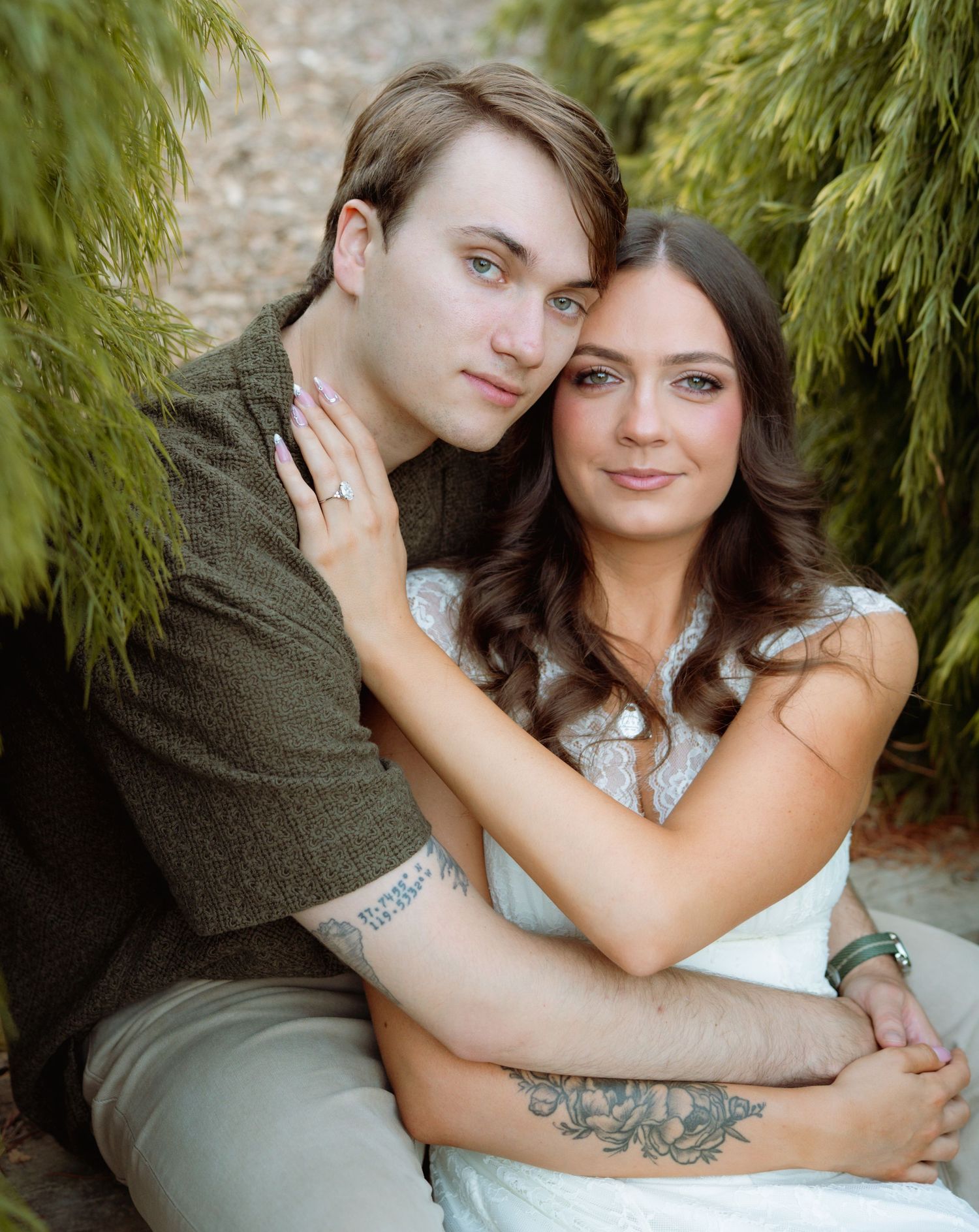 A young couple embraces tenderly among green weeping trees, the woman showing a ring and tattoos.