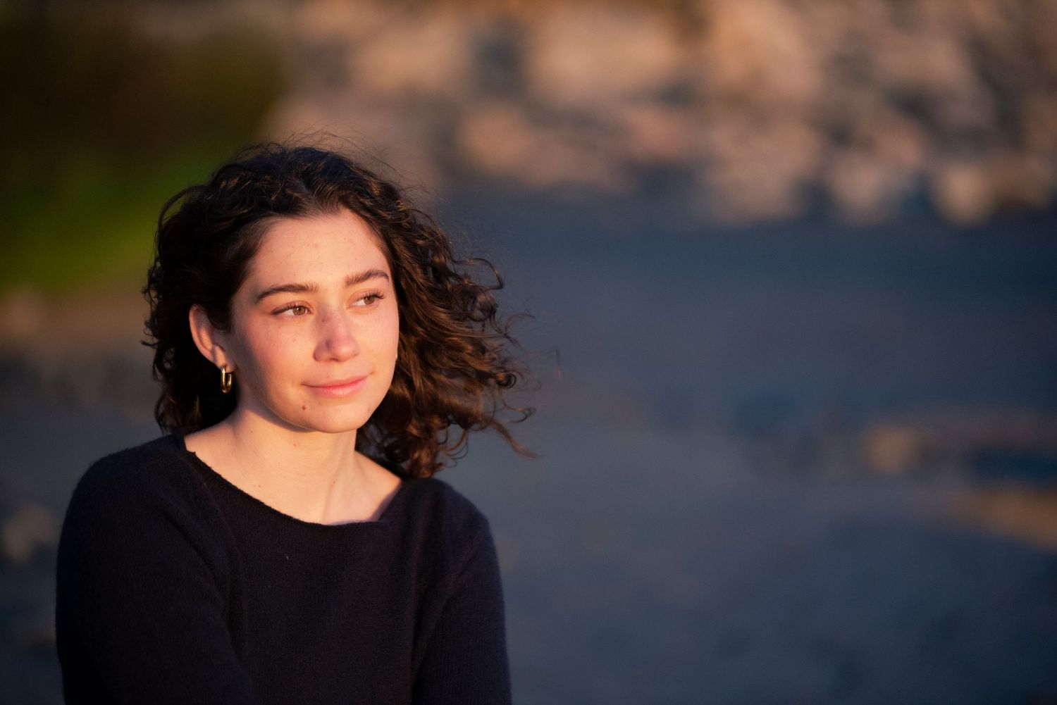 Young woman with curly hair smiling softly outdoors in warm golden hour light.
