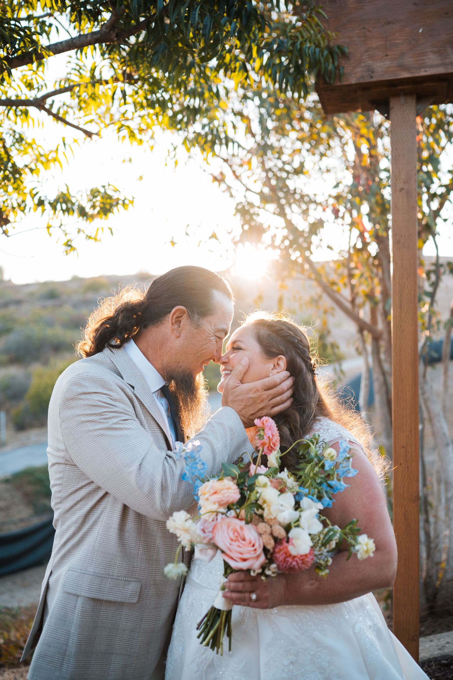 Groom tenderly cups brides face as they lean in for a kiss with golden sunlight glowing behind them.