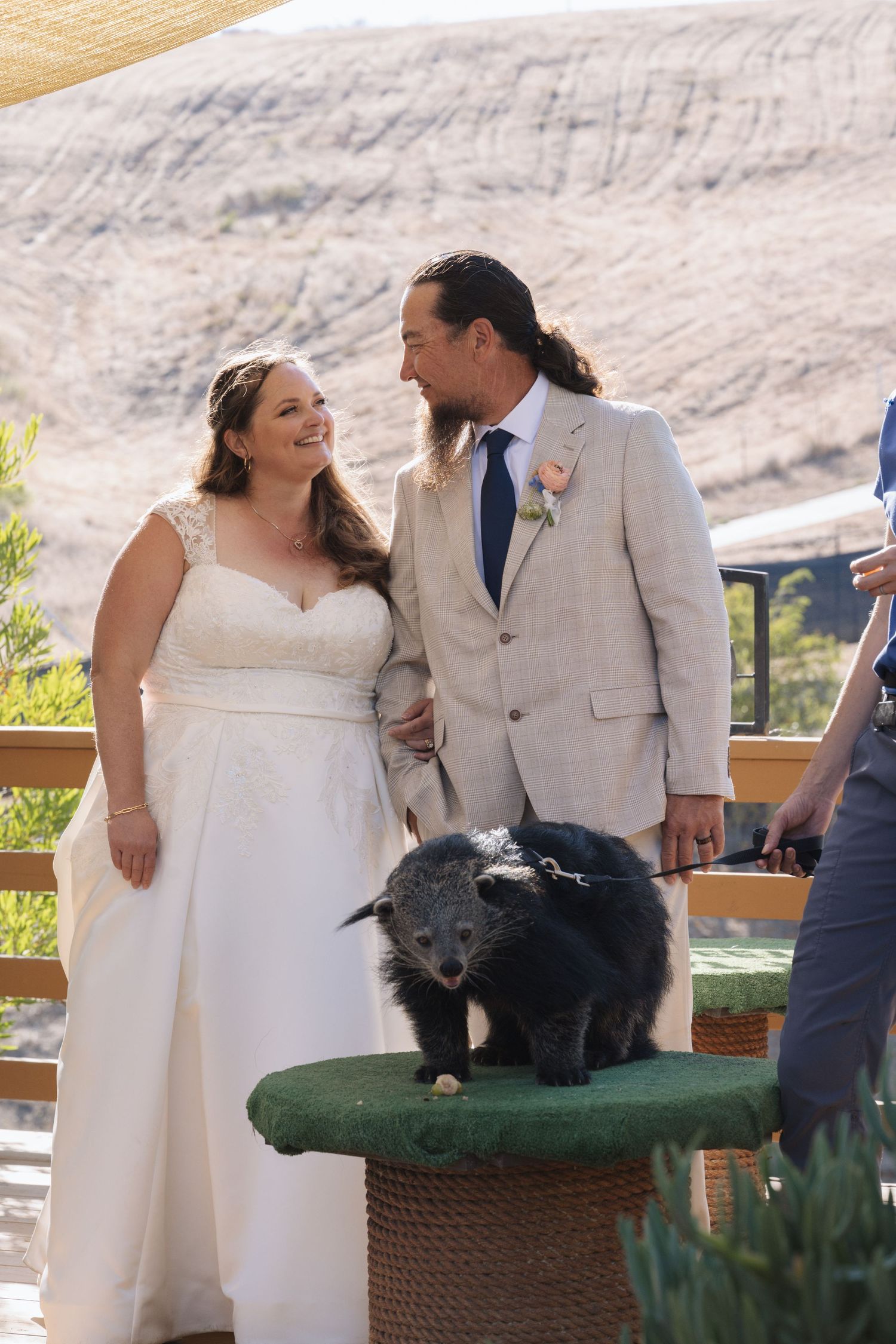 Bride and groom smile at each other during outdoor wedding ceremony with a small black bear on a pedestal beside them.