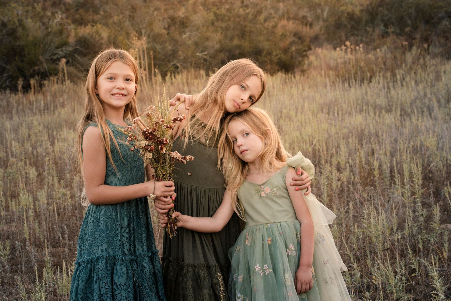 Three young girls in teal dresses pose together in a golden meadow holding wildflowers.