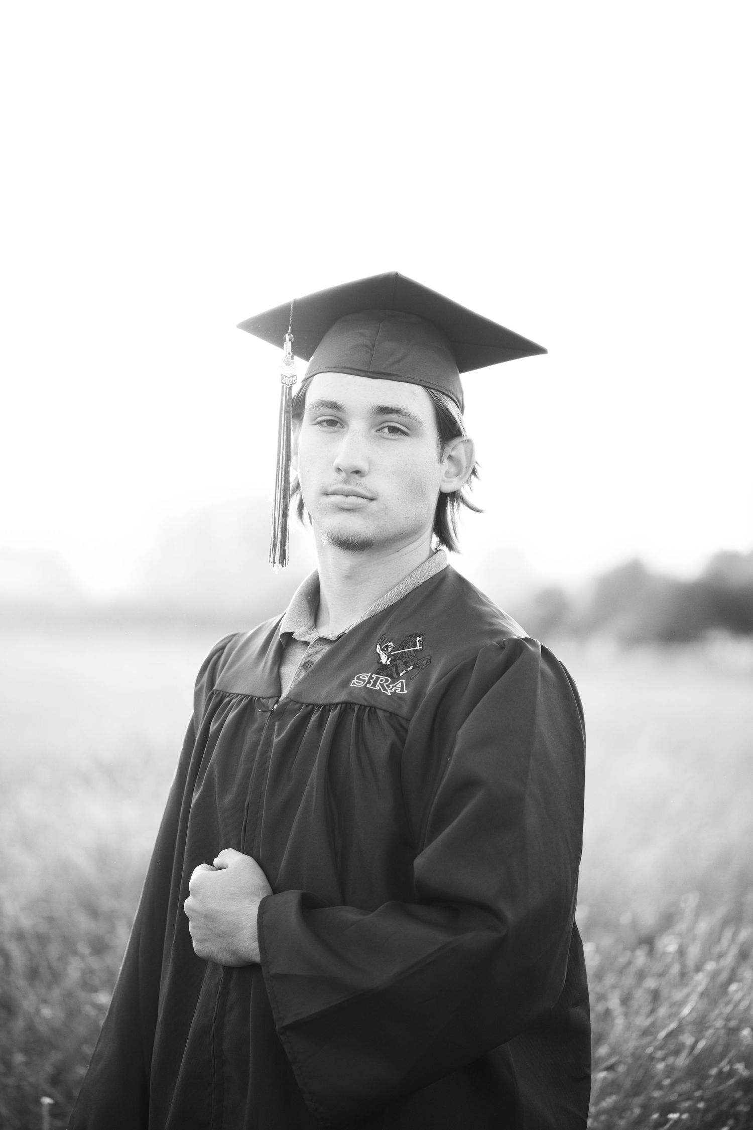 Black and white portrait of graduate in cap and gown standing in outdoor field at dusk.