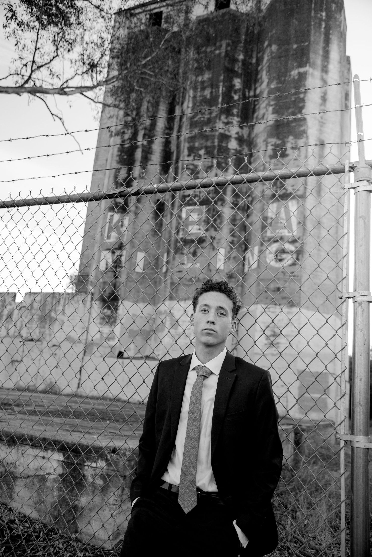 Young man in black suit leaning against chain-link fence near abandoned building in black and white photo.