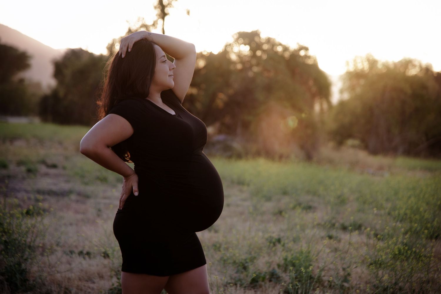 Pregnant woman in black dress standing in grassy field during golden hour sunset.